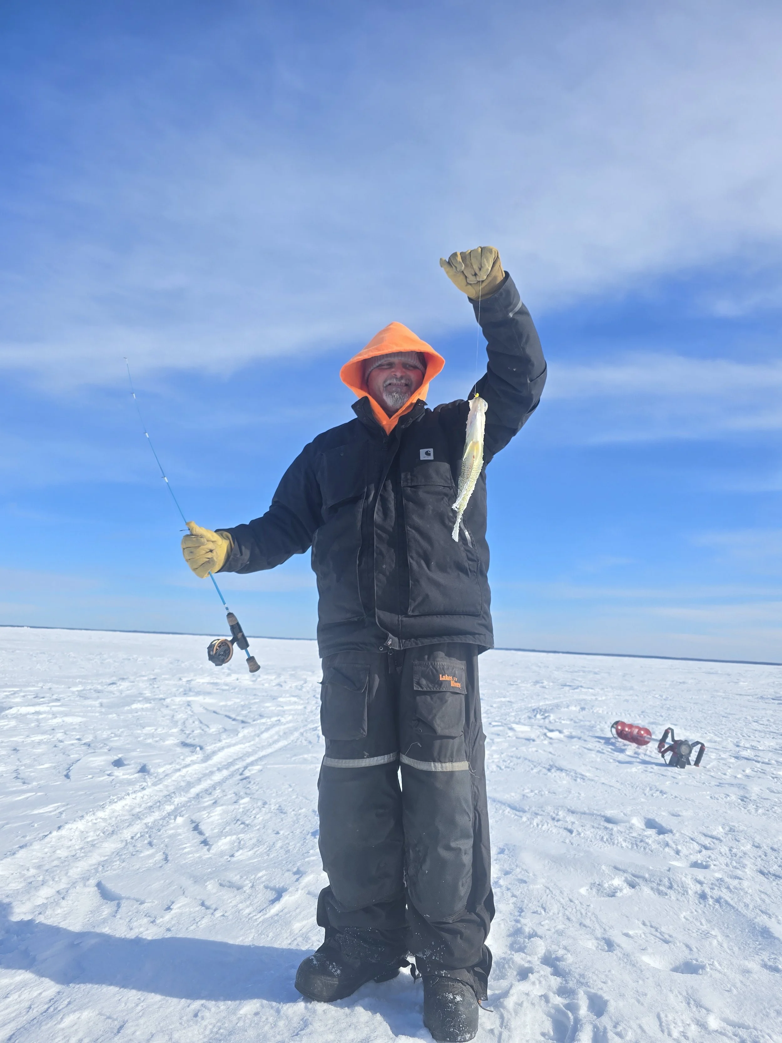 A man in winter clothing holding up a fish on a line, standing in a snowy landscape under a blue sky.