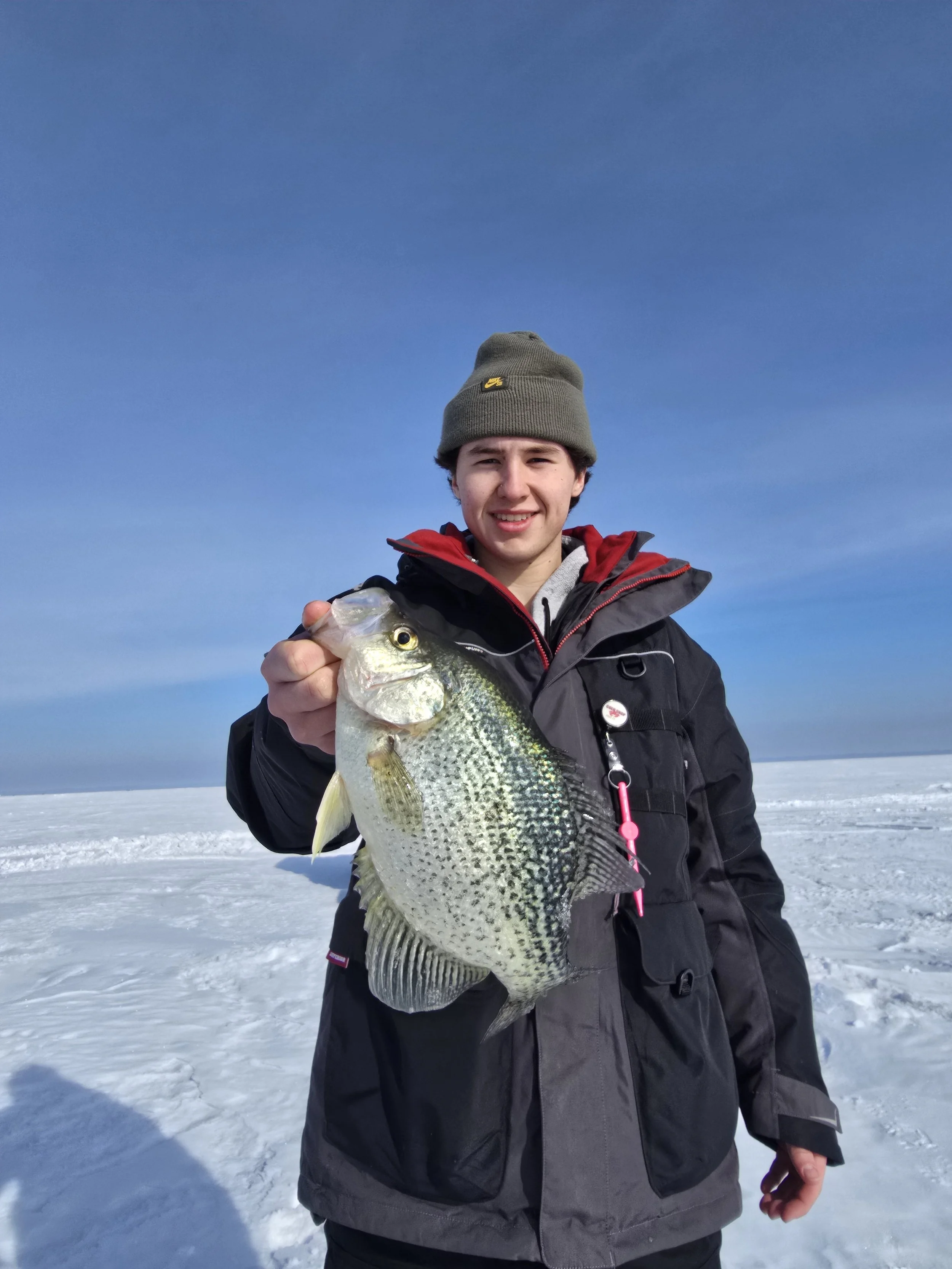 A young man standing on snowy ground holding a large fish, wearing a gray beanie and a black winter jacket with a red lining, with a clear blue sky in the background.