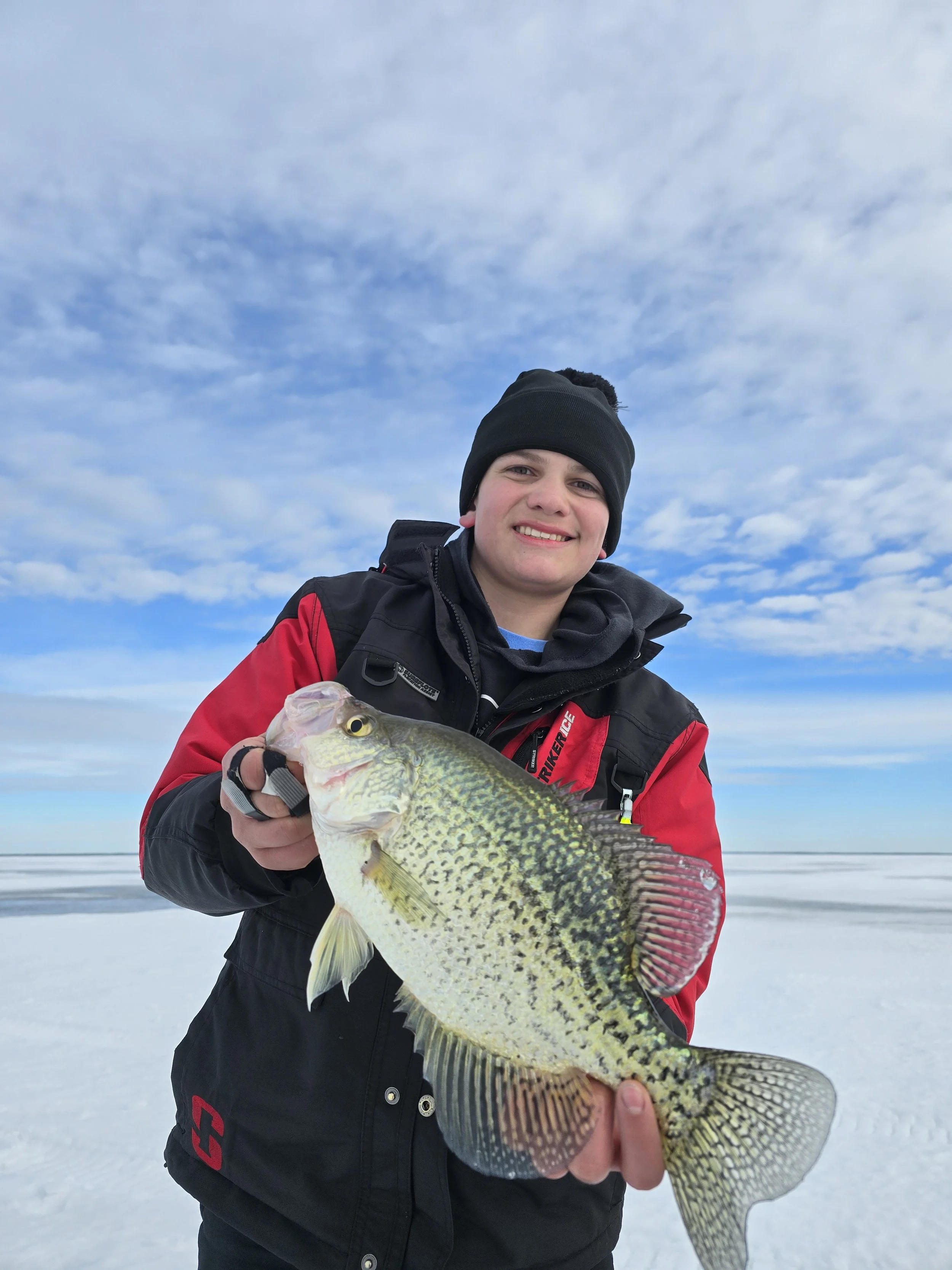 A young person dressed in a black winter hat and a red and black jacket stands outdoors on a snowy landscape, holding a large fish with yellow and black markings, under a cloudy sky.