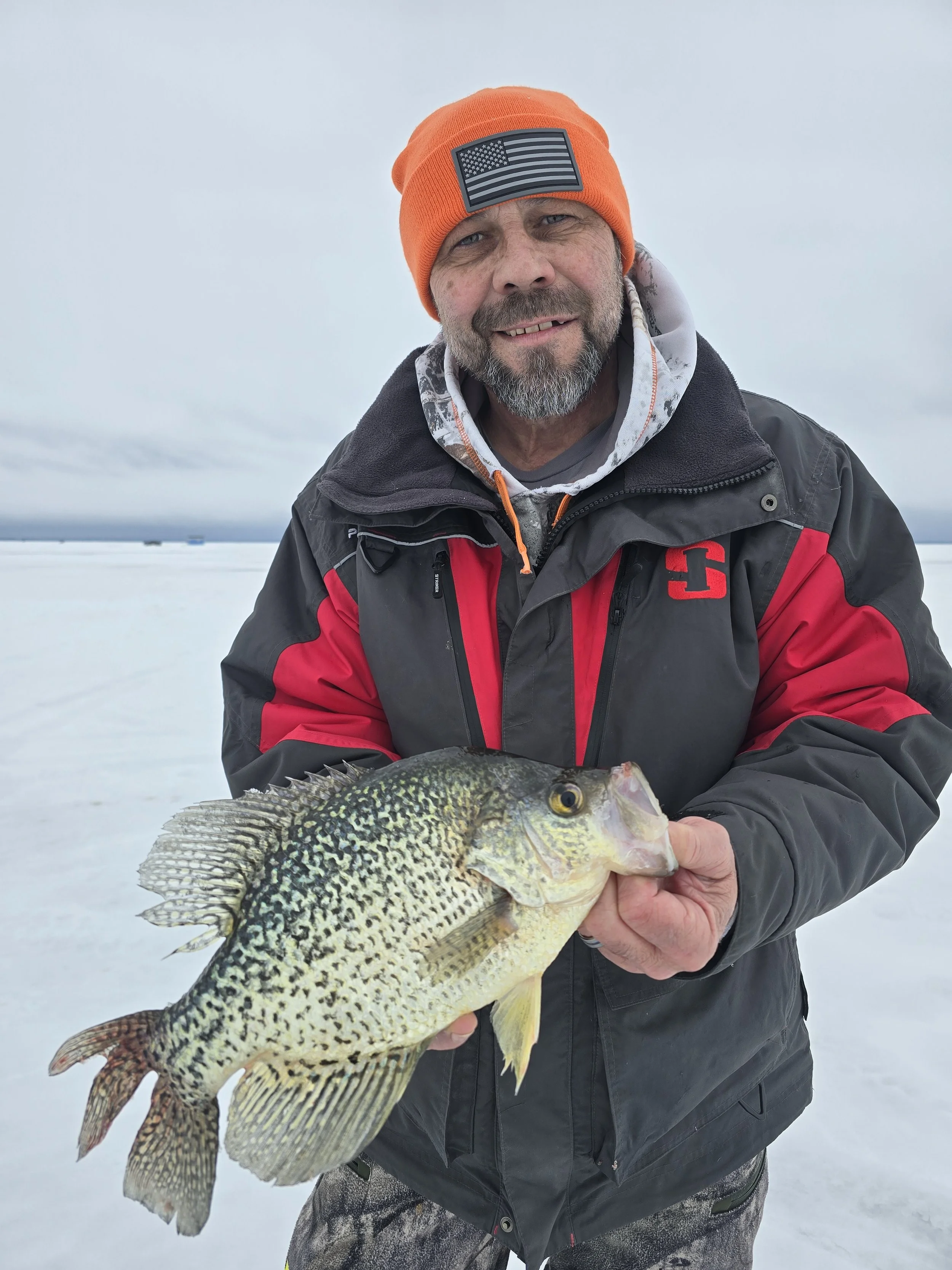 Man holding a large fish on snowy landscape, wearing orange beanie and winter jacket.
