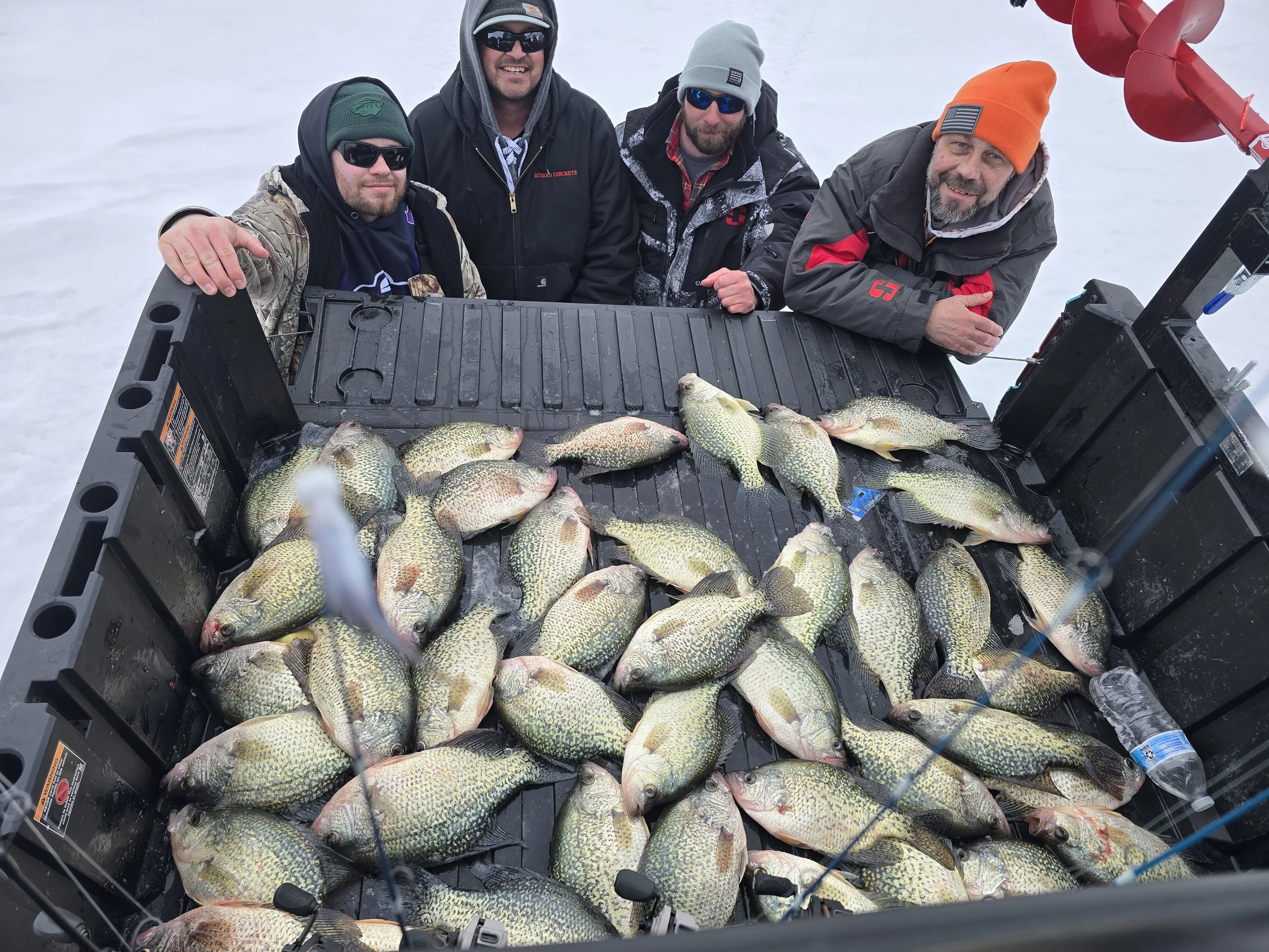 Four men dressed in winter clothing and sunglasses pose behind a large catch of fish laid out on a black plastic surface, with snow in the background and a red ice auger on the right side.