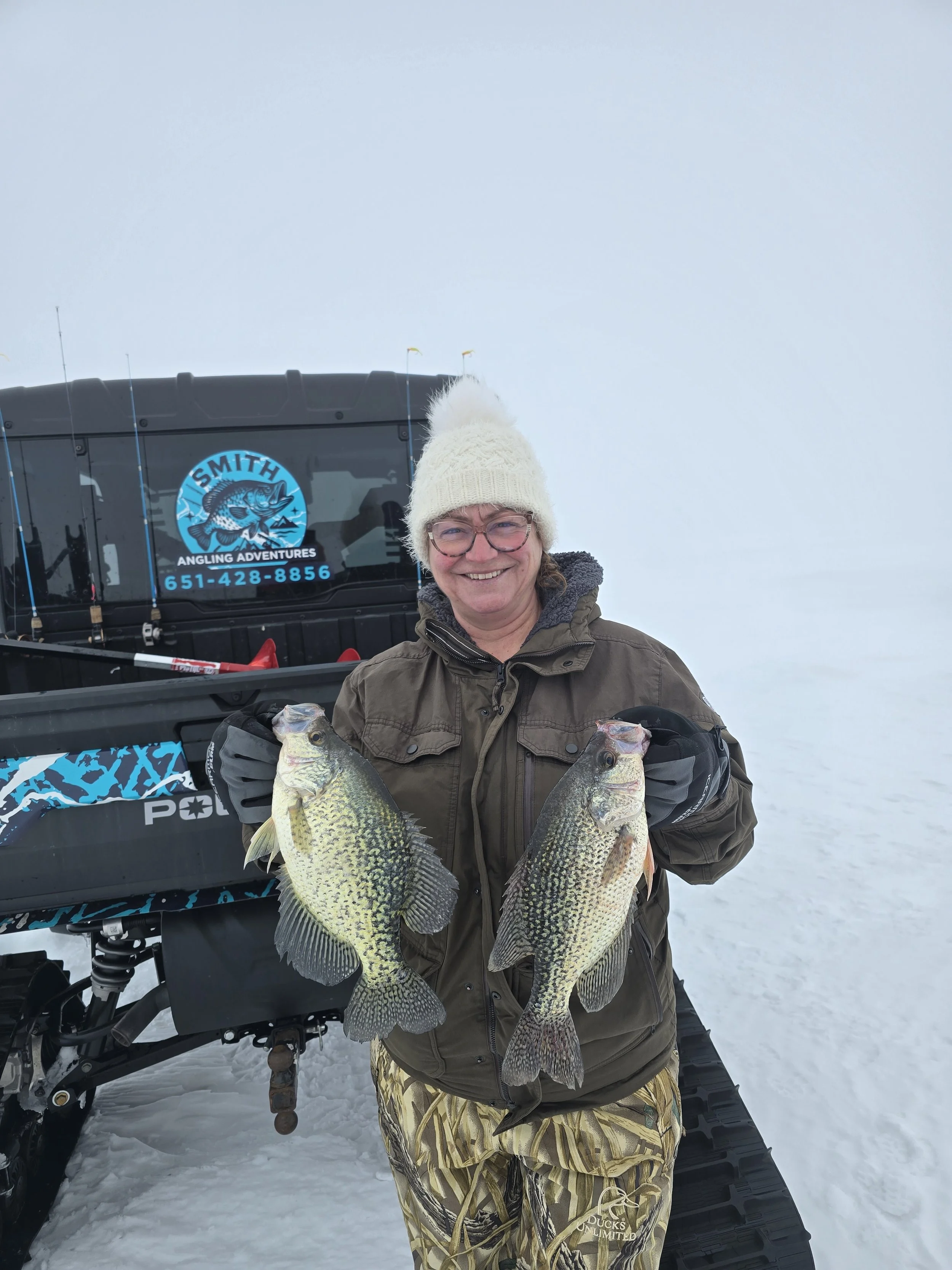 Woman smiling and holding two fish in front, standing on snow, with a snowmobile and fishing gear in background.