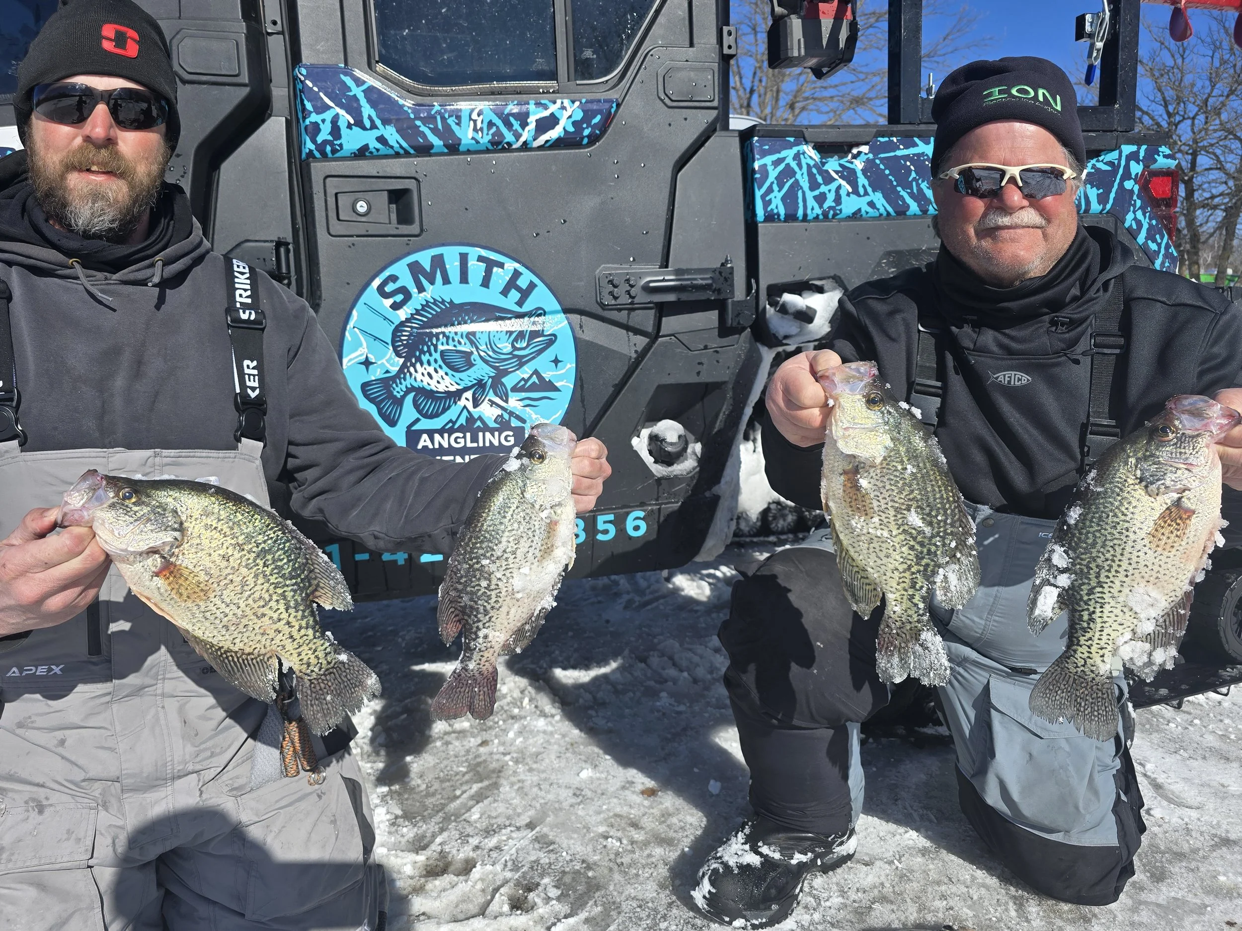 Two men holding large fish, posing in front of a snow-covered outdoor setting with a vehicle in the background. Both are wearing winter clothing and sunglasses.