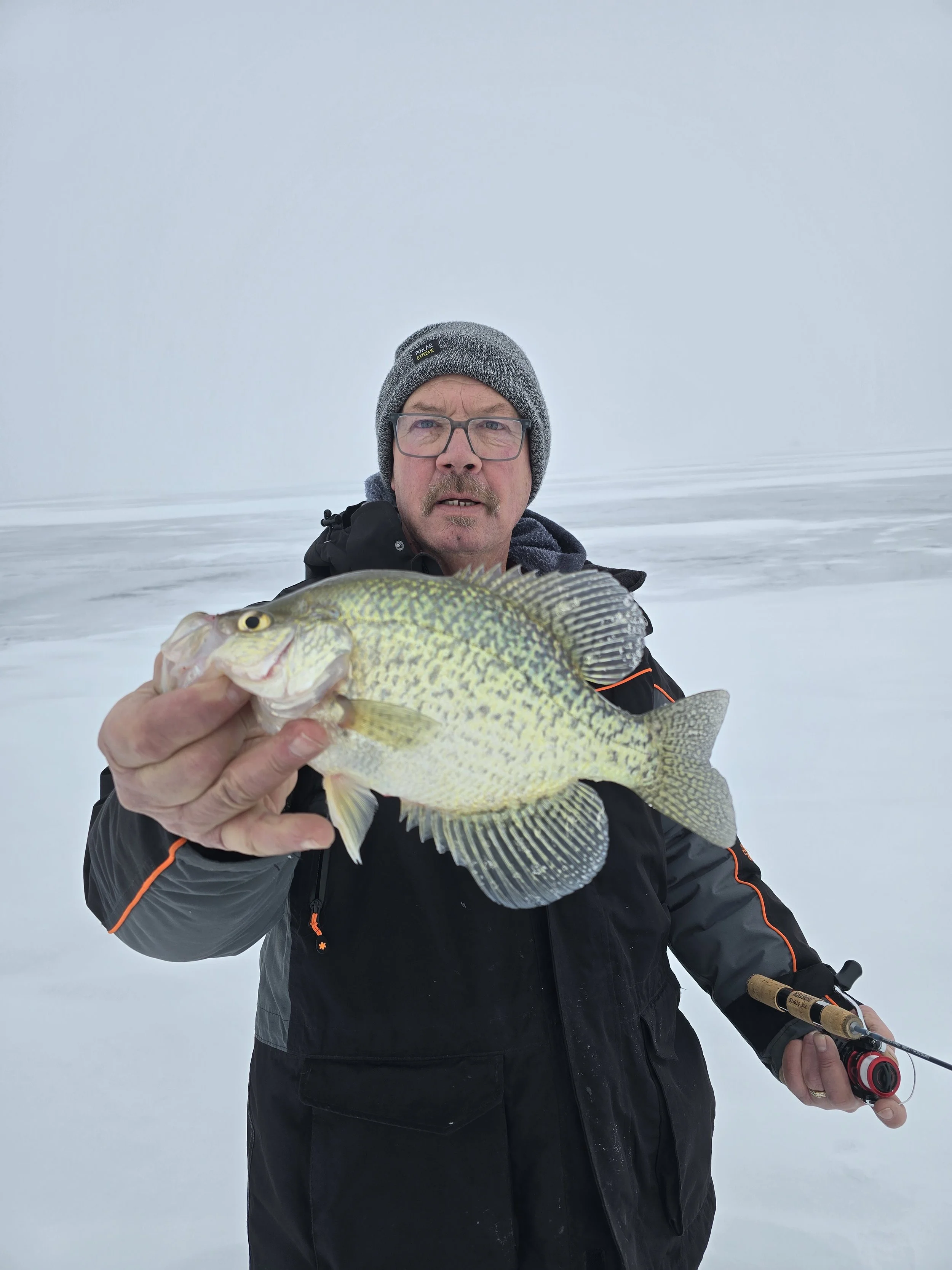 A man in a gray beanie, glasses, black jacket with orange accents, holding a fishing rod in one hand and a fish in the other, standing in a snowy landscape under an overcast sky.
