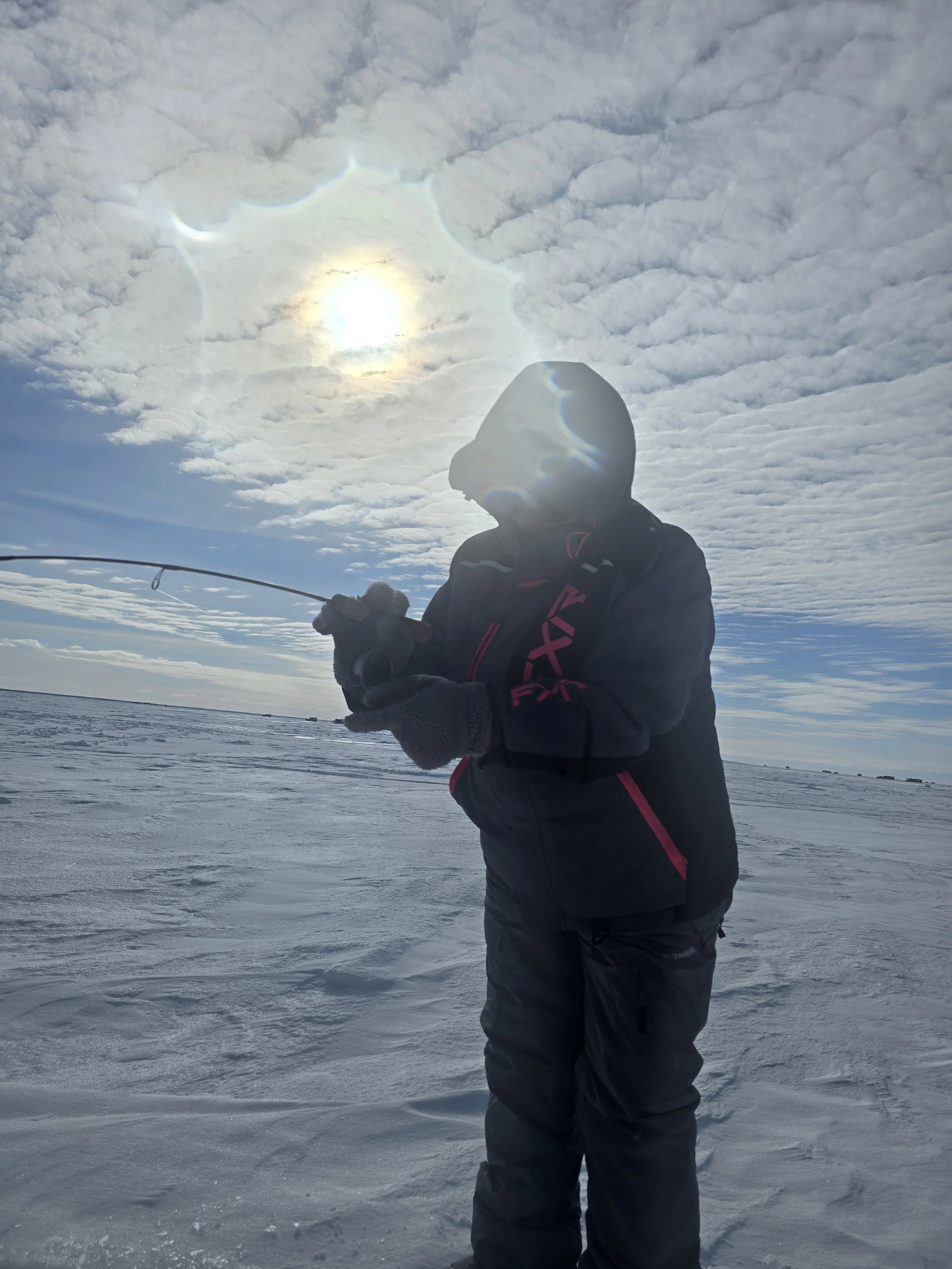 A person dressed in winter clothing is ice fishing on a frozen body of water with a fishing rod, under a cloudy sky with the sun partially obscured.