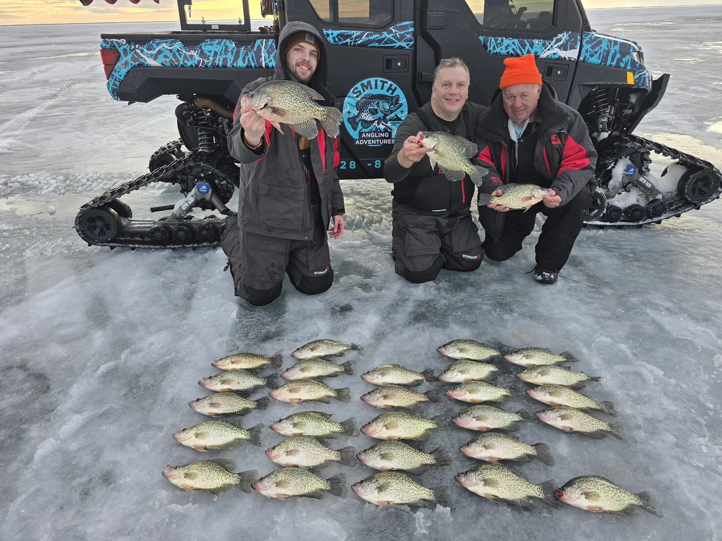Three men kneeling on ice in front of a snowmobile, holding fish, with fish lined up on the ice in front of them and a logo on the snowmobile that says 'Smith Angling Adventures.'