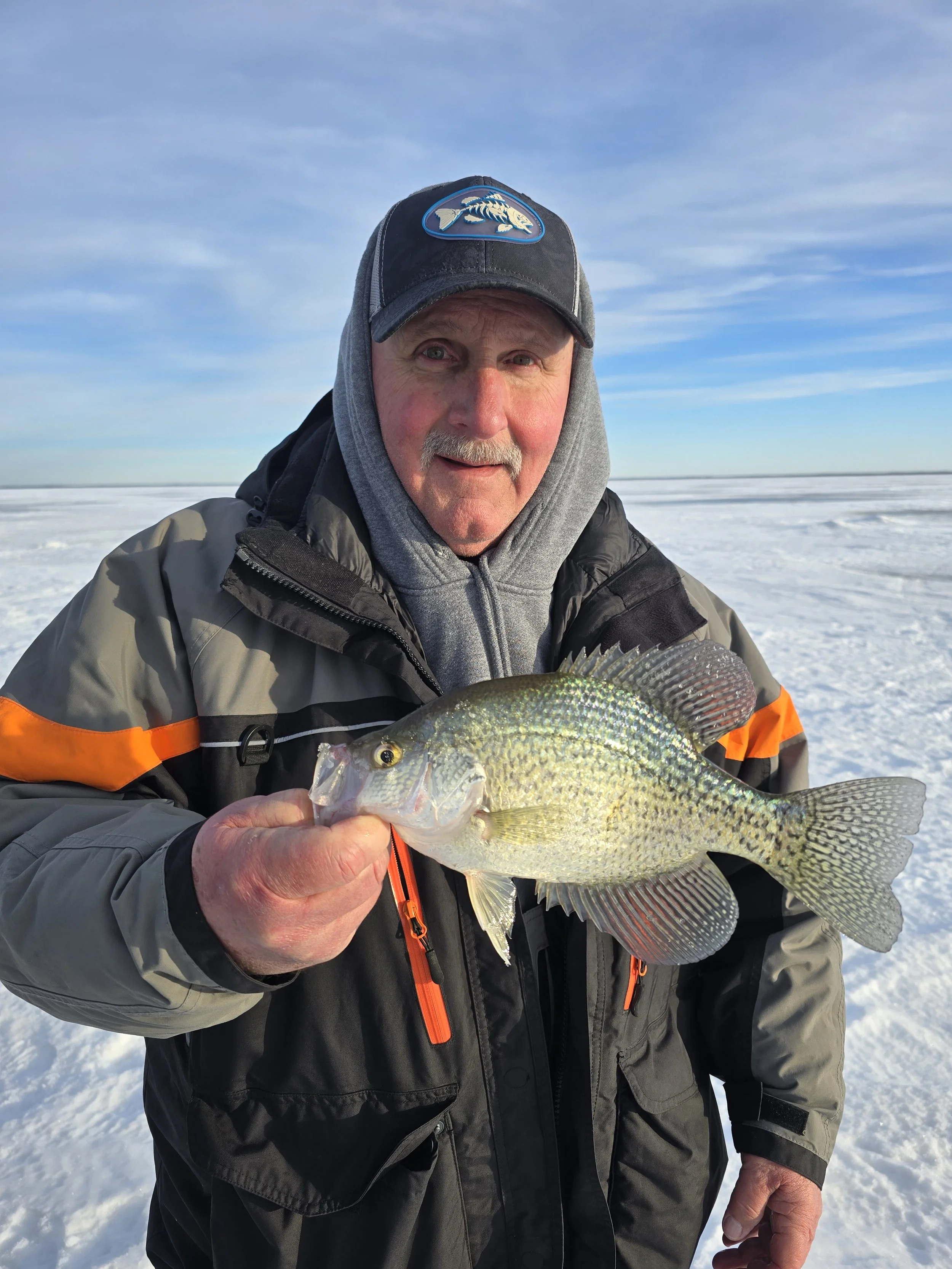 A man dressed in winter clothing holding a fish on a snowy landscape with a clear blue sky in the background.