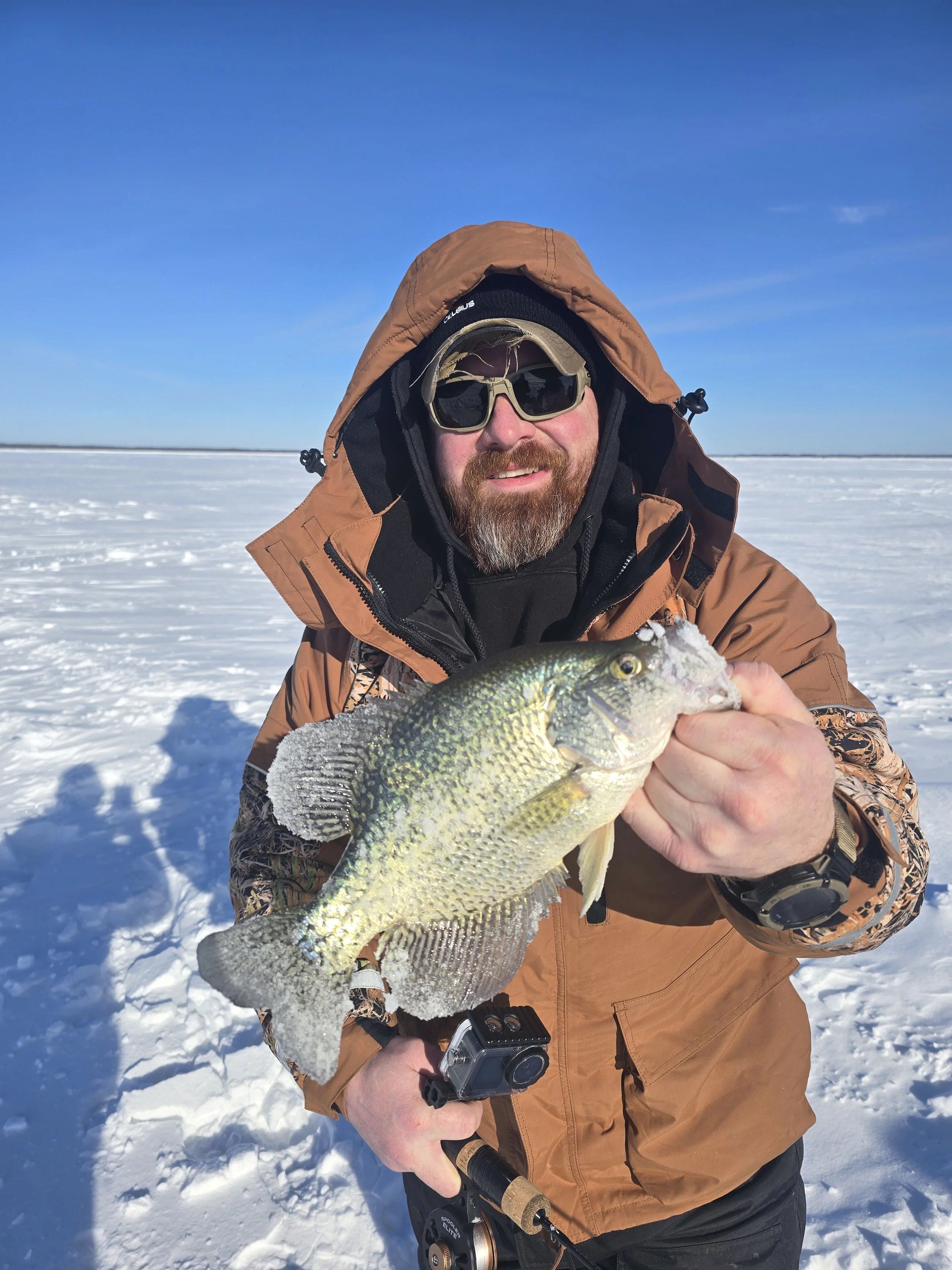 A man in winter clothing, sunglasses, and a cap smiling and posing with a fish he caught over a snowy landscape with a clear blue sky.