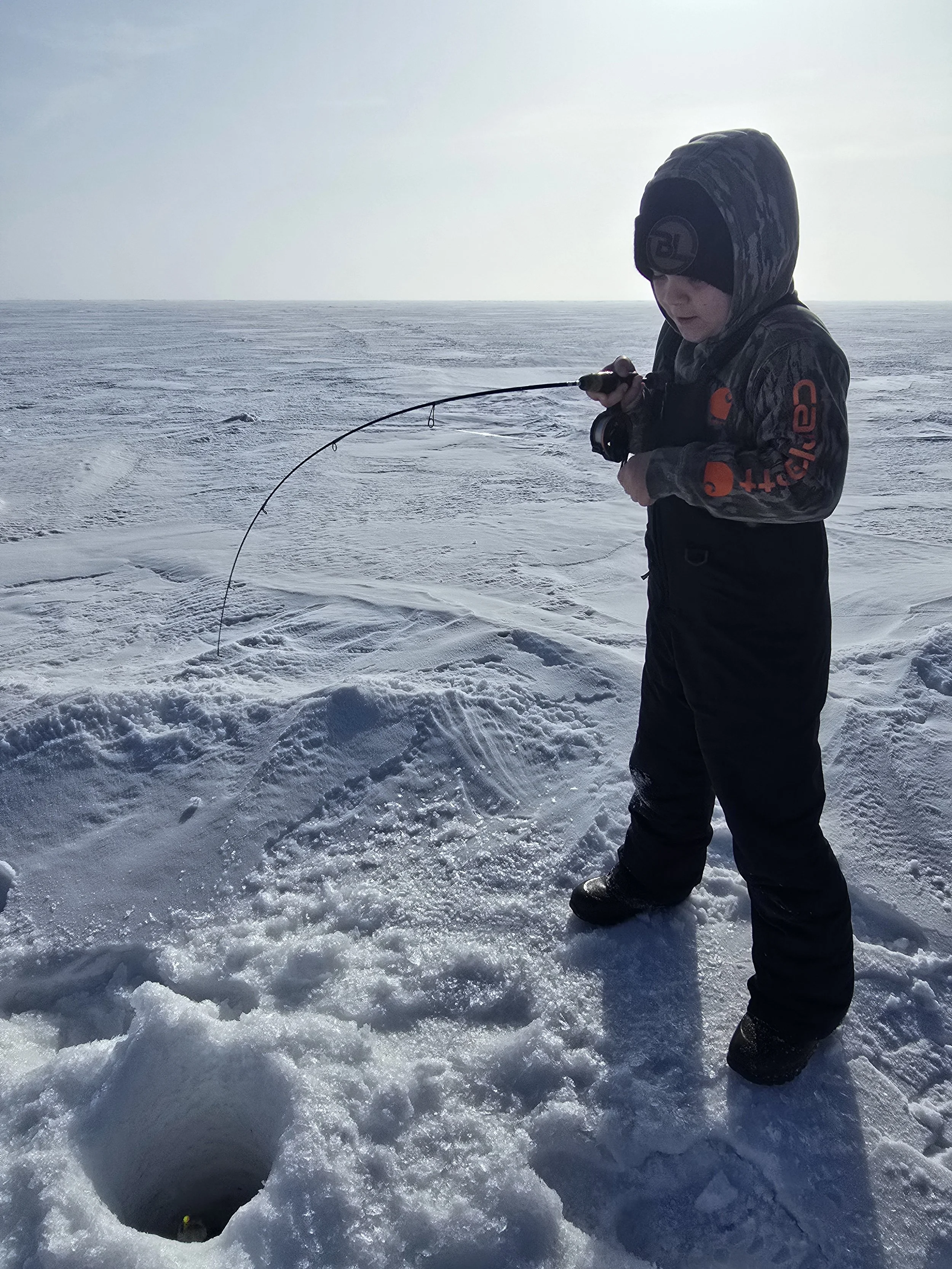 Child ice fishing on snowy landscape, holding a fishing rod, wearing winter gear and a hood.