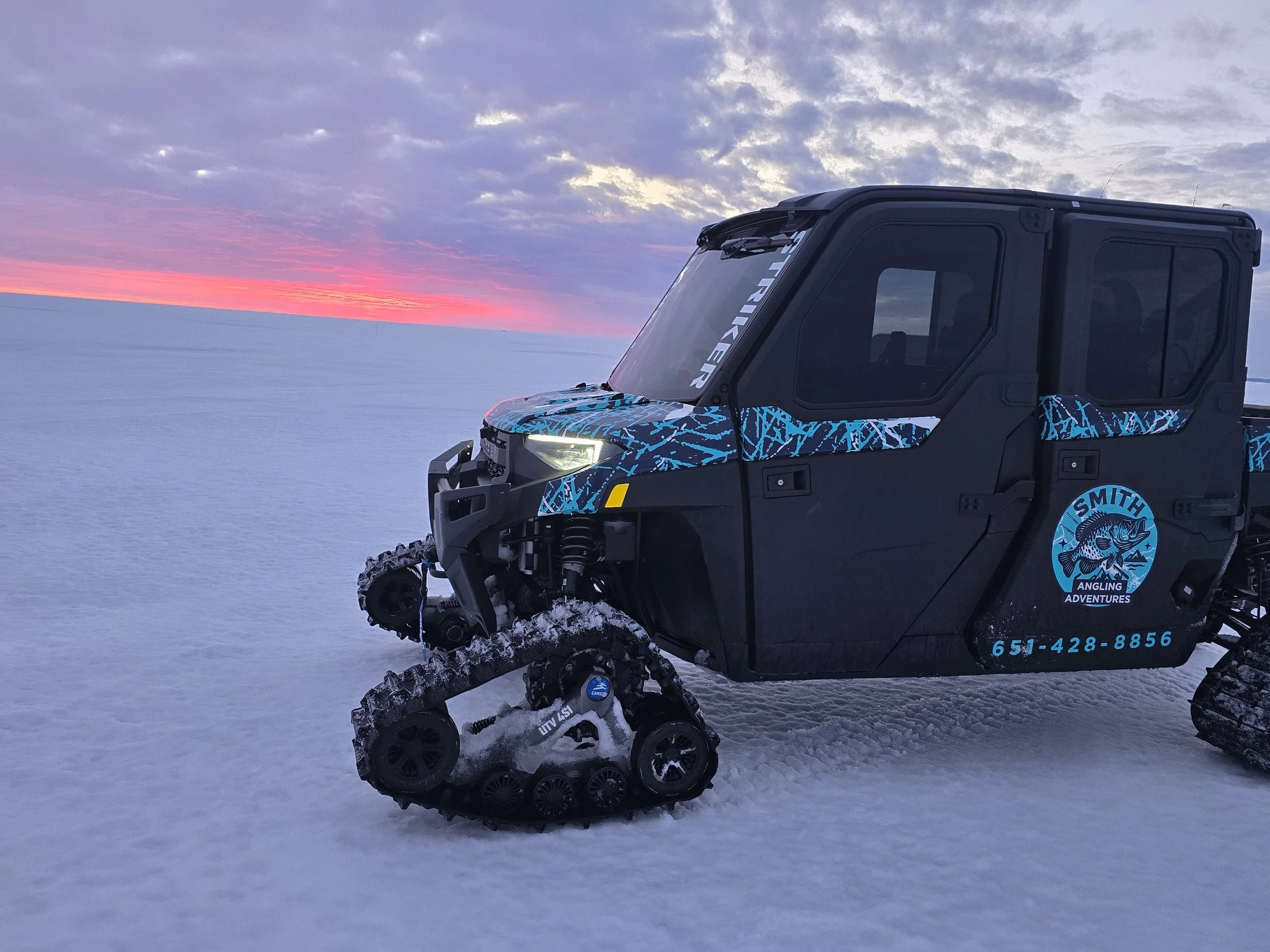 An all-terrain vehicle (ATV) on snow with a sunset sky in the background. The ATV has a blue and black design with a logo that says "Fishing Adventures."