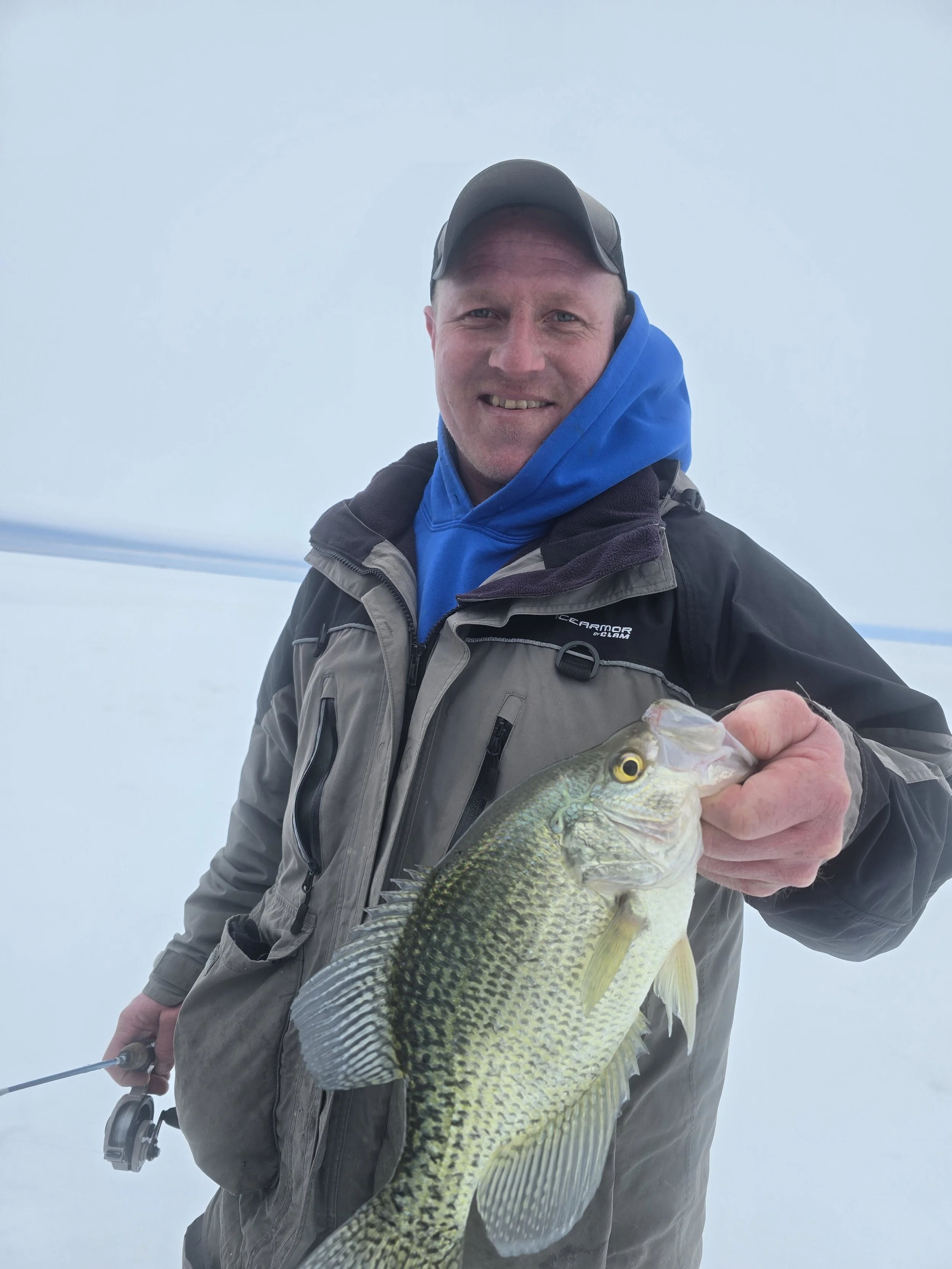 A man wearing a gray jacket, a blue hoodie, and a gray cap, holding a large fish with green and yellow scales in his right hand. He is smiling, and the background is foggy or cloudy with faint power lines visible.