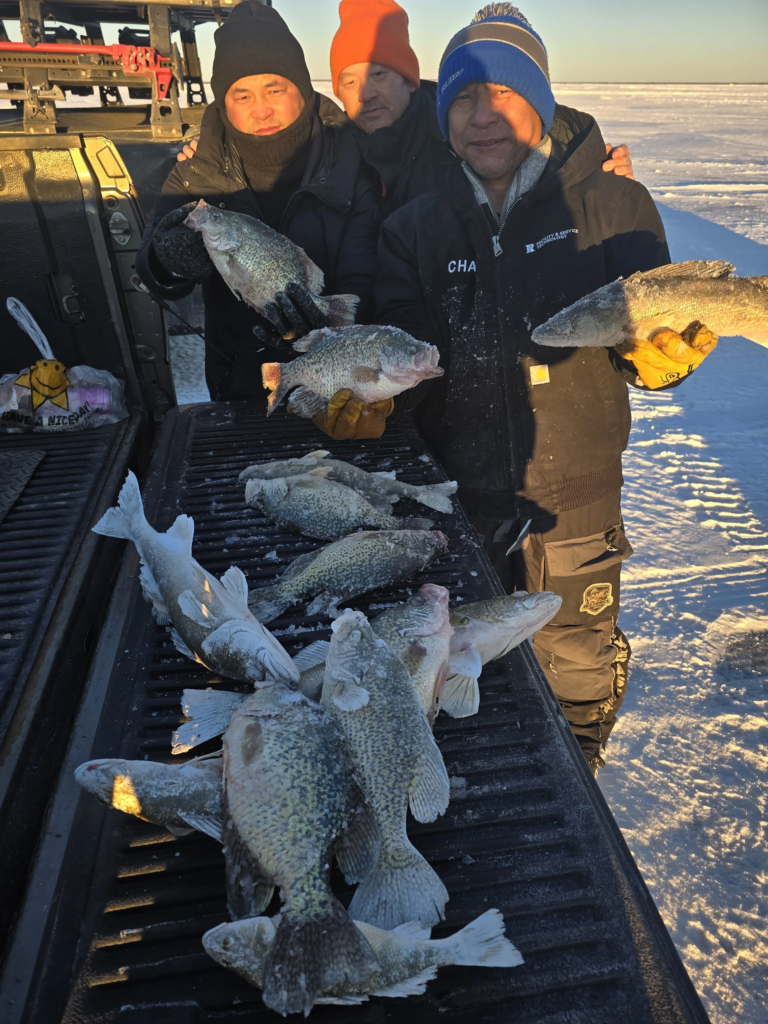 Three men in winter clothing display multiple fish caught through ice fishing on a frozen lake during sunset.