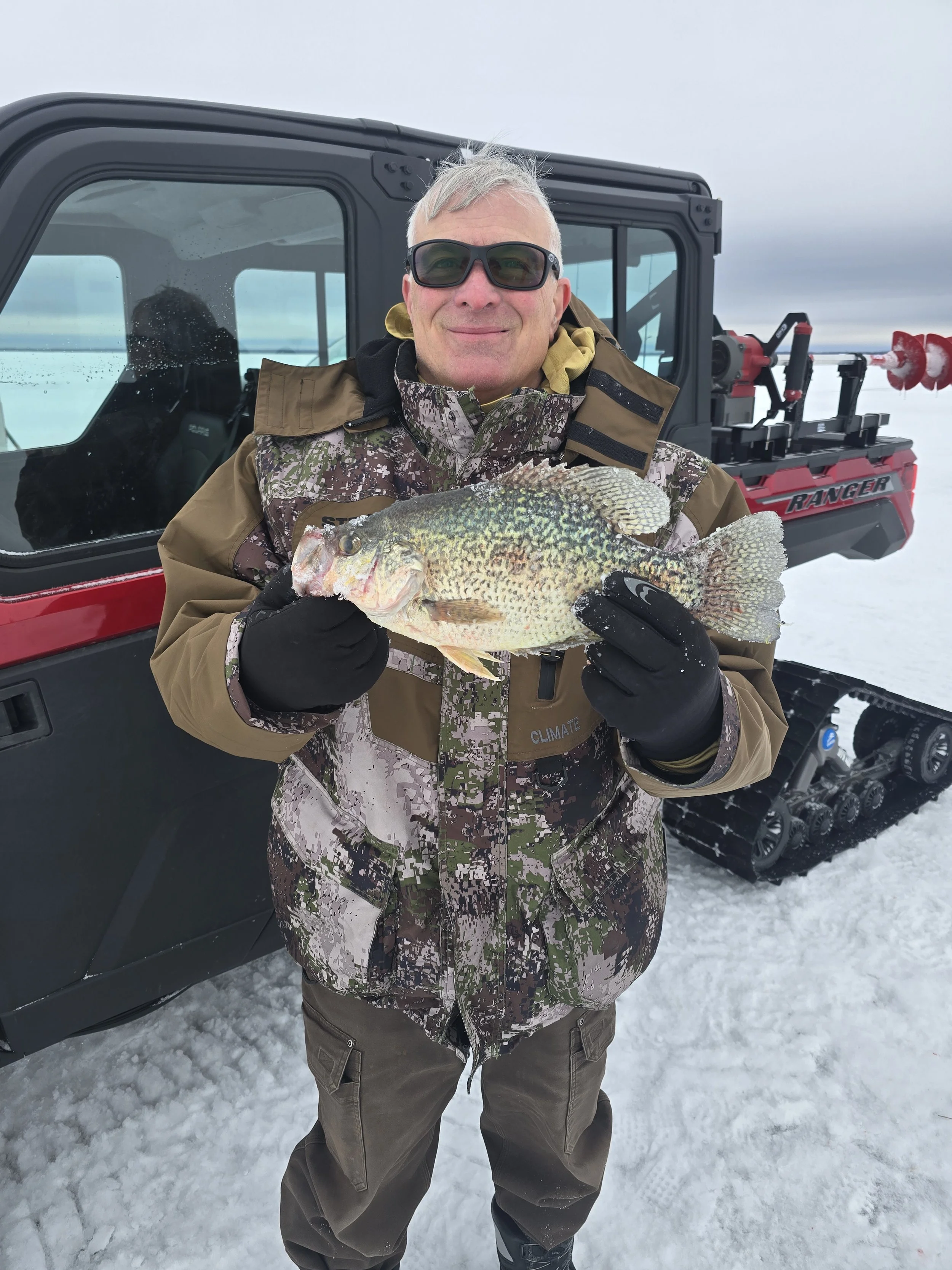 Man holding a large fish on snow, wearing camouflage jacket, black gloves, sunglasses, and brown pants, with snowmobiles and overcast sky in background.
