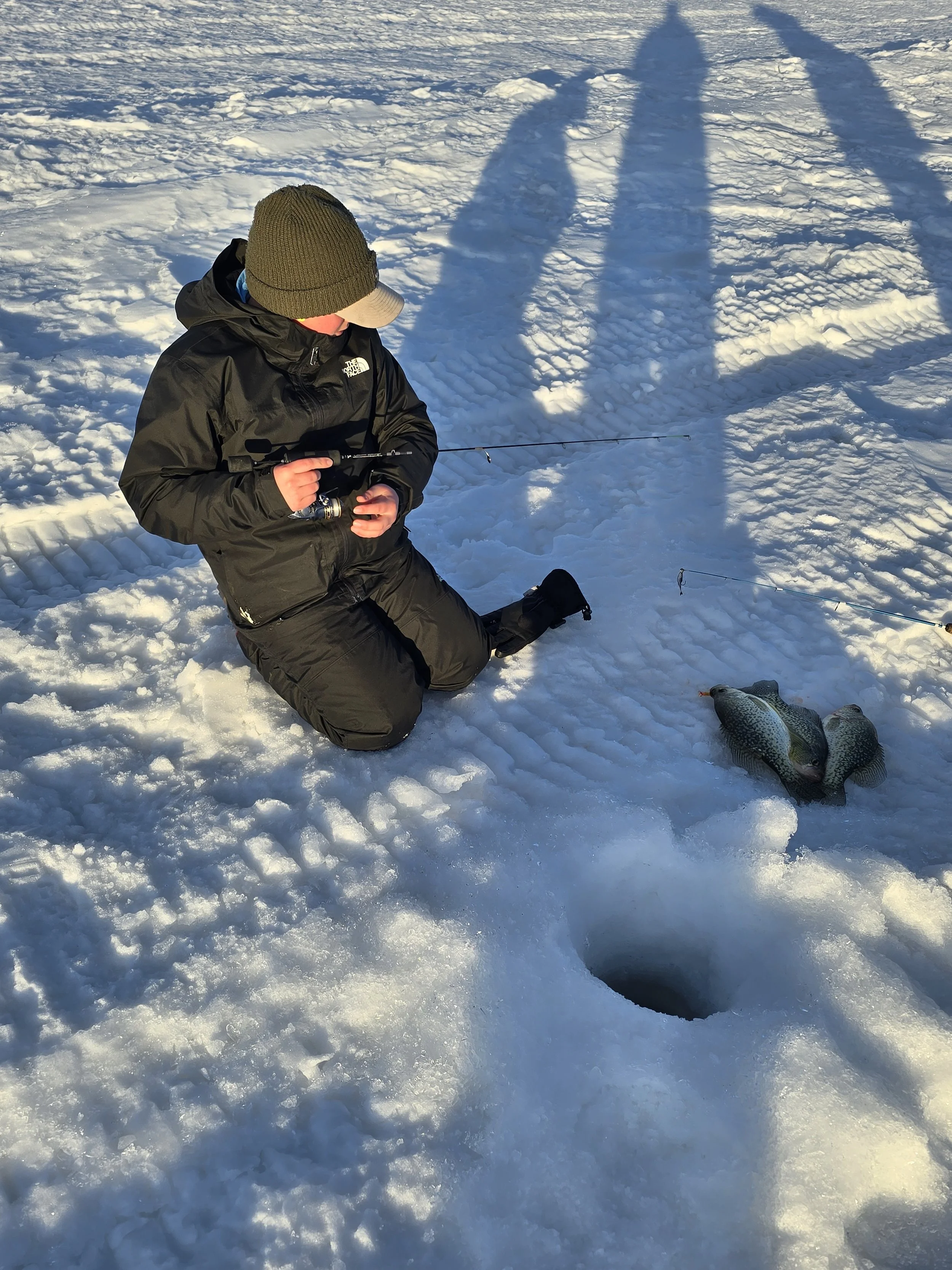 A person kneeling on snow with a fishing rod, two large fish caught and lying on the snow, casting a shadow on the snow in bright sunlight.