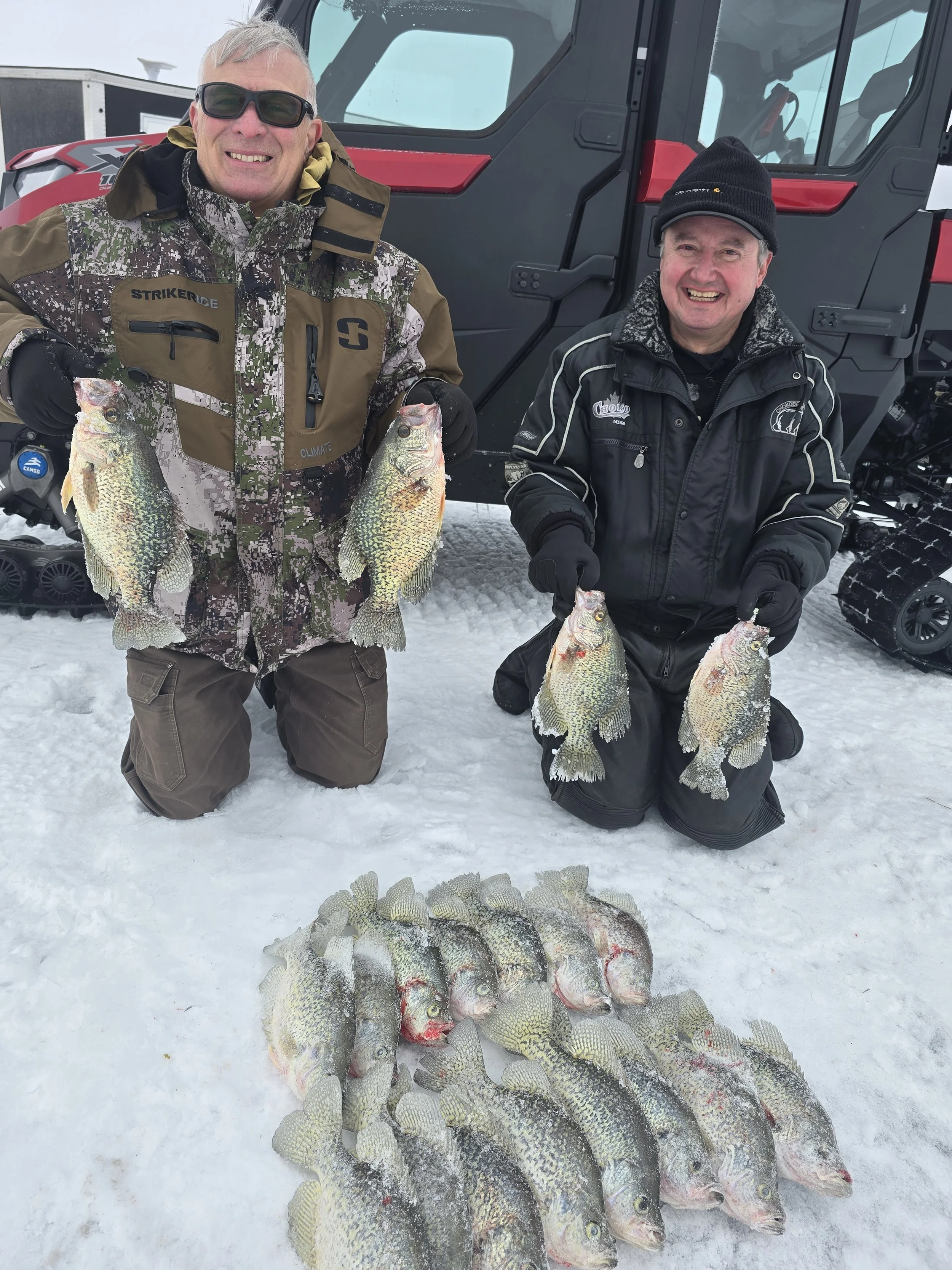 Two men kneeling on snow in front of a large vehicle, displaying crappie fish they caught. The man on the left is wearing camouflage clothing and sunglasses, holding two fish, while the man on the right is wearing a black jacket and beanie, holding t