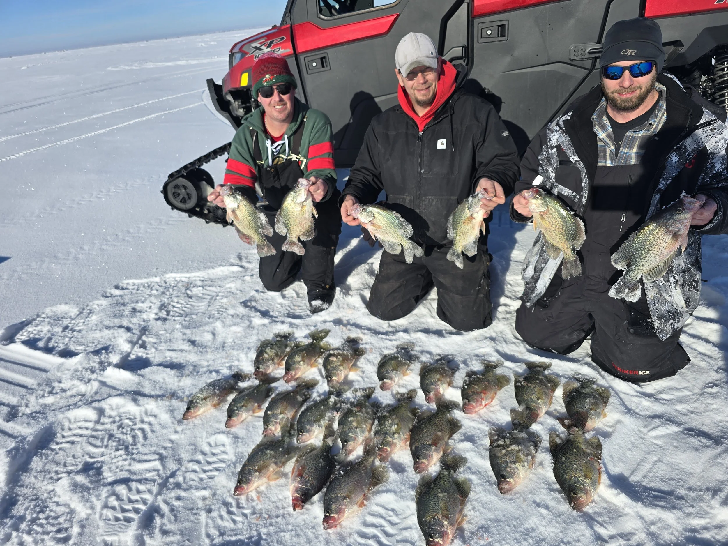 Three men are kneeling on snow-covered ice, each holding multiple fish they caught, with a large vehicle in the background.