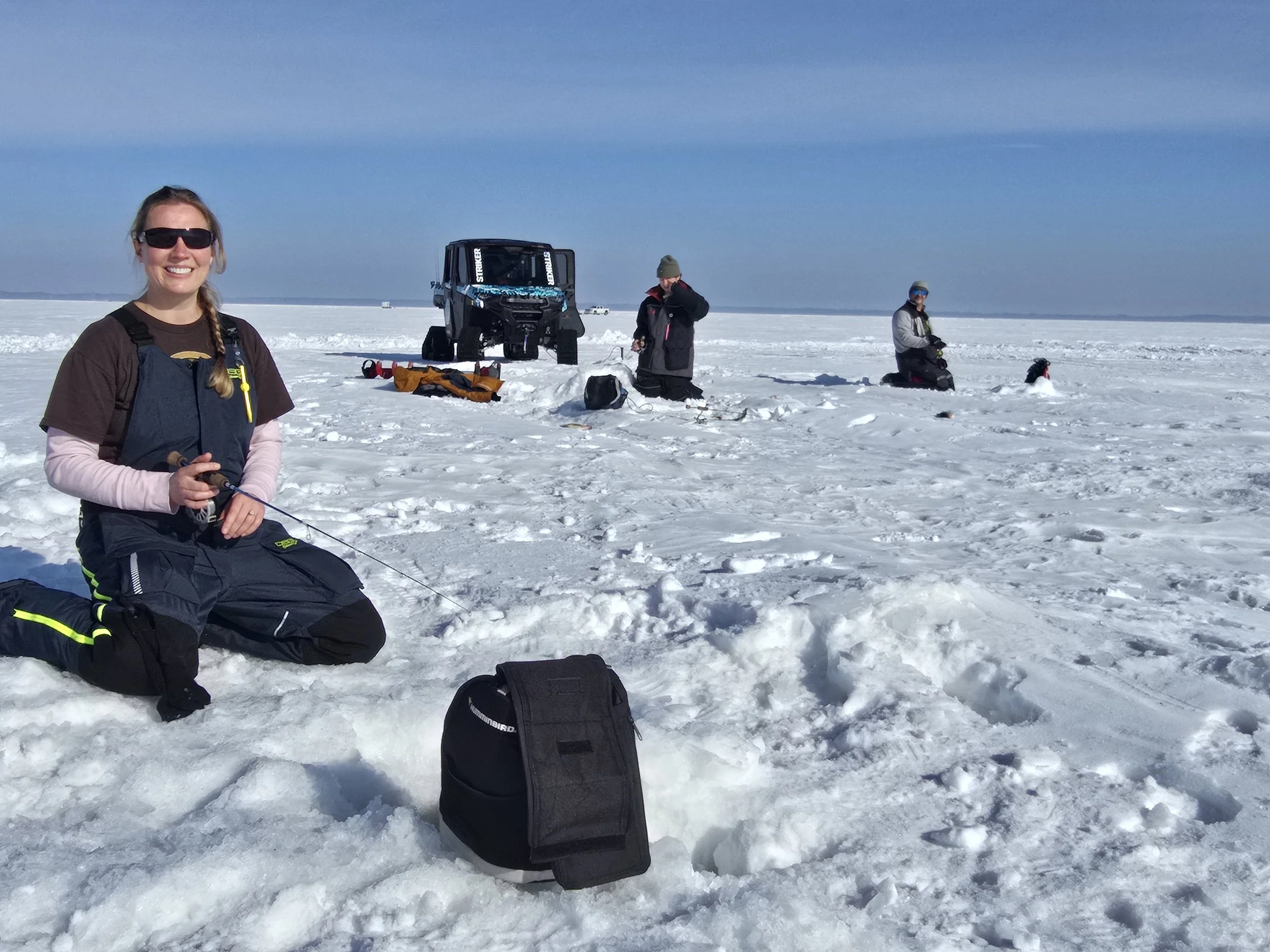 Four people in winter clothing with gear on a snowy, frozen landscape under a clear blue sky, engaging in an outdoor activity possibly ice fishing or research.
