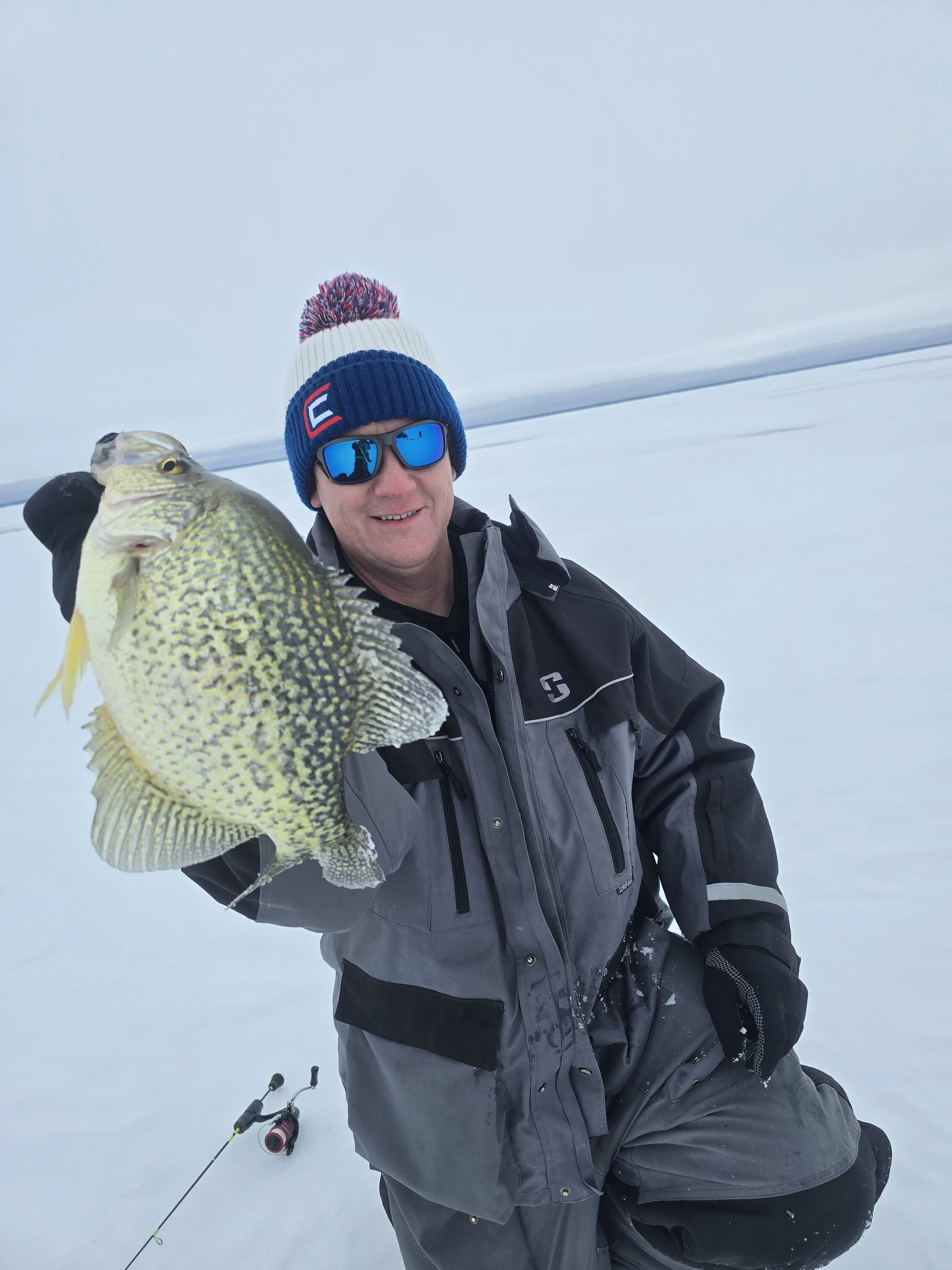 Man in winter clothing holding a large fish on snowy landscape with ice fishing gear nearby.