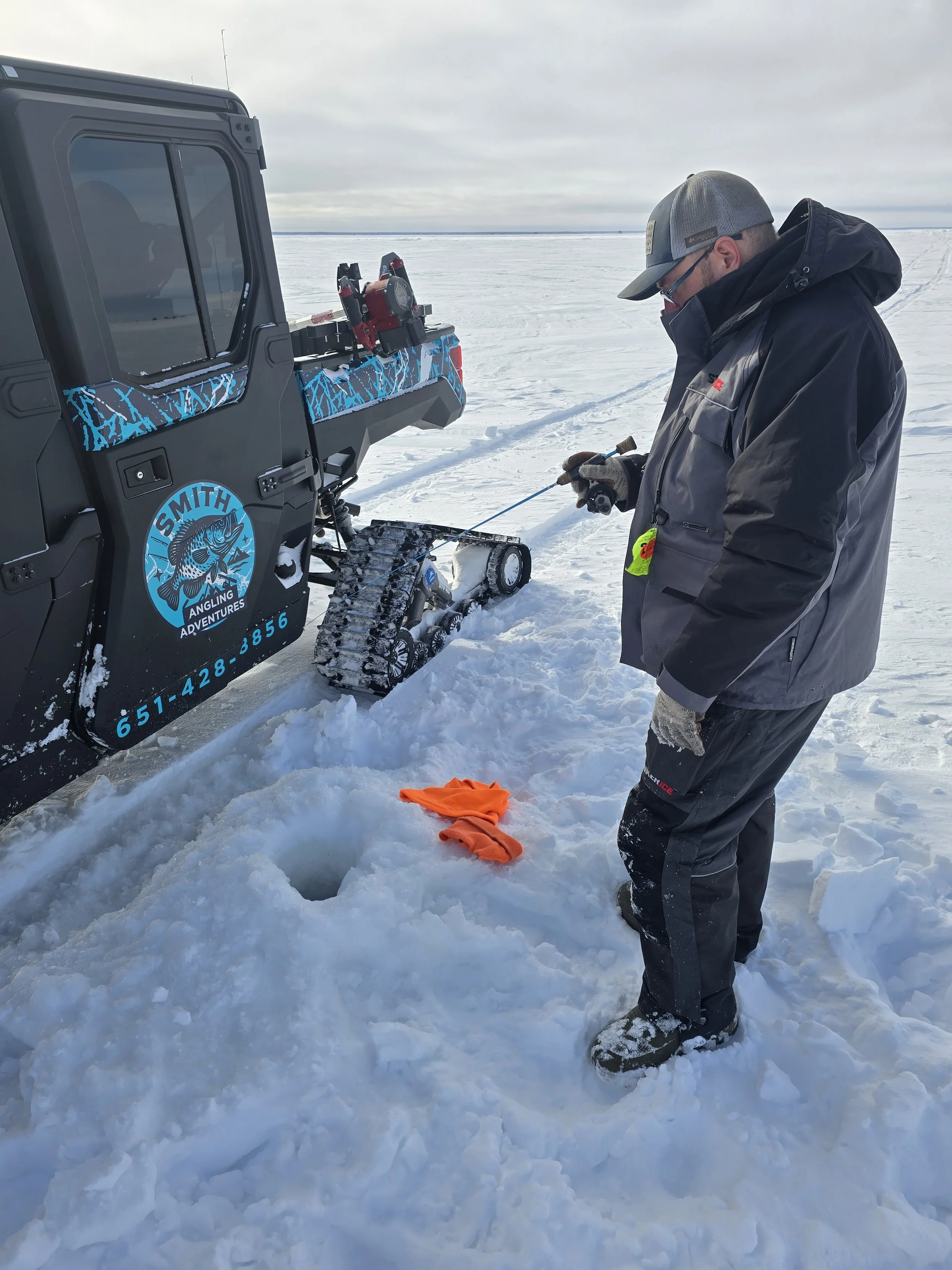 A man in winter gear stands on snow-covered ice, holding a small tool, near an arctic research vehicle with a logo that says 'Smith Angling Adventures' and a fish illustration.