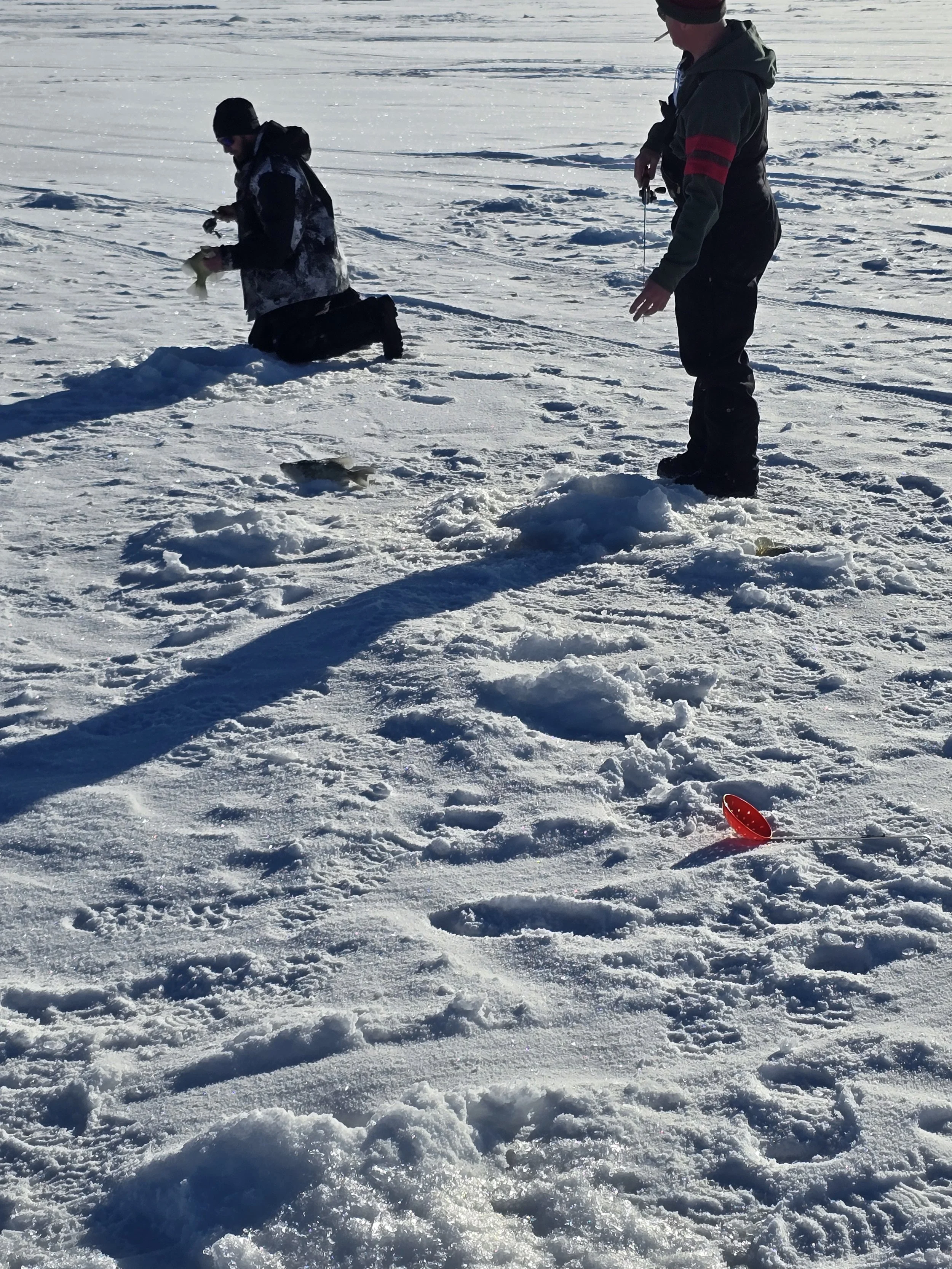 Two men ice fishing on a snowy frozen lake; one is kneeling with a fishing line, the other stands holding a fishing rod, with a small red fishing container on the snow nearby.