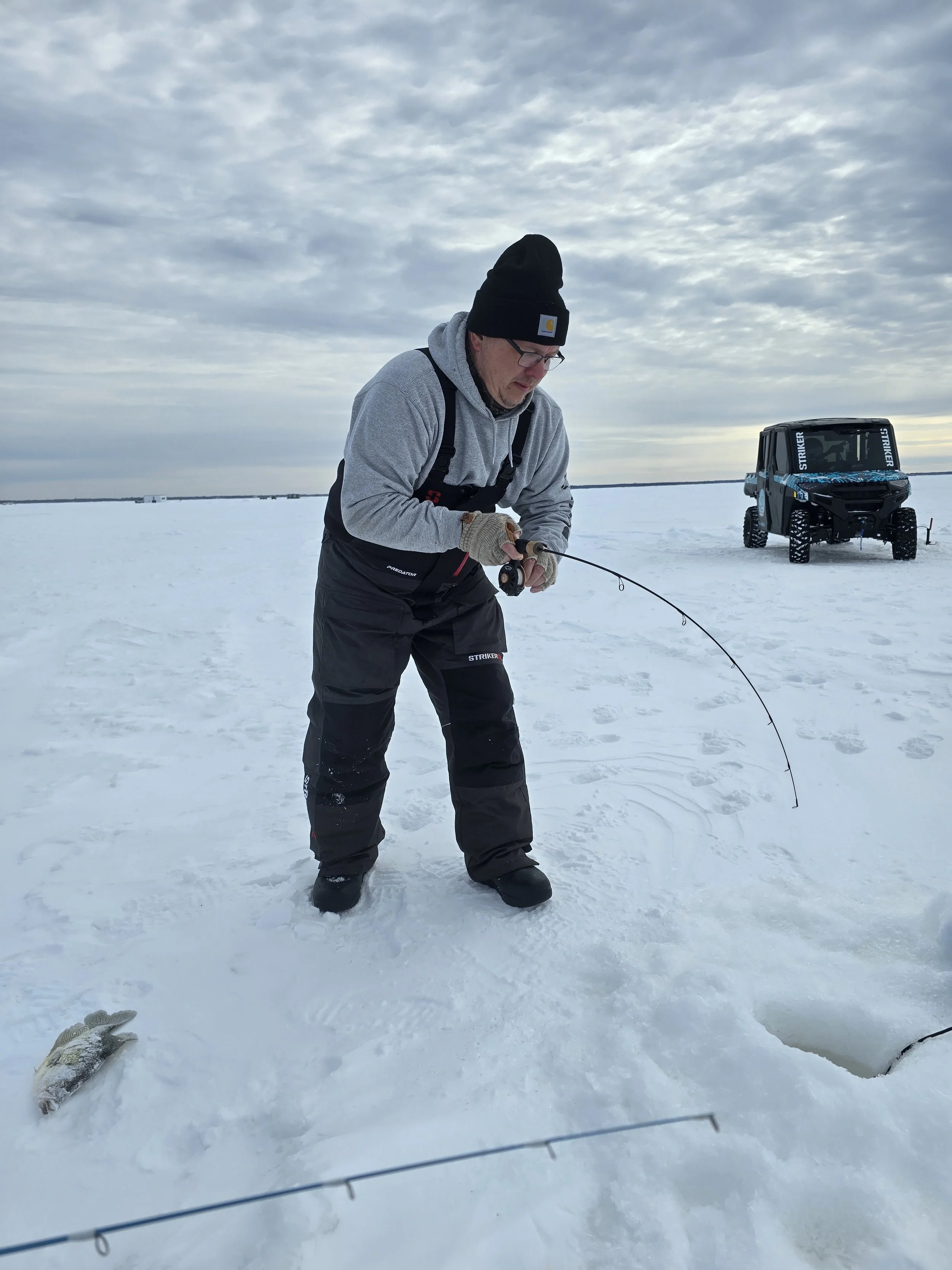 A man in winter clothing, wearing a gray hoodie, black beanie, and gloves, is ice fishing on a snow-covered frozen lake. There is a black utility vehicle in the background on the ice. The sky is overcast with gray clouds.