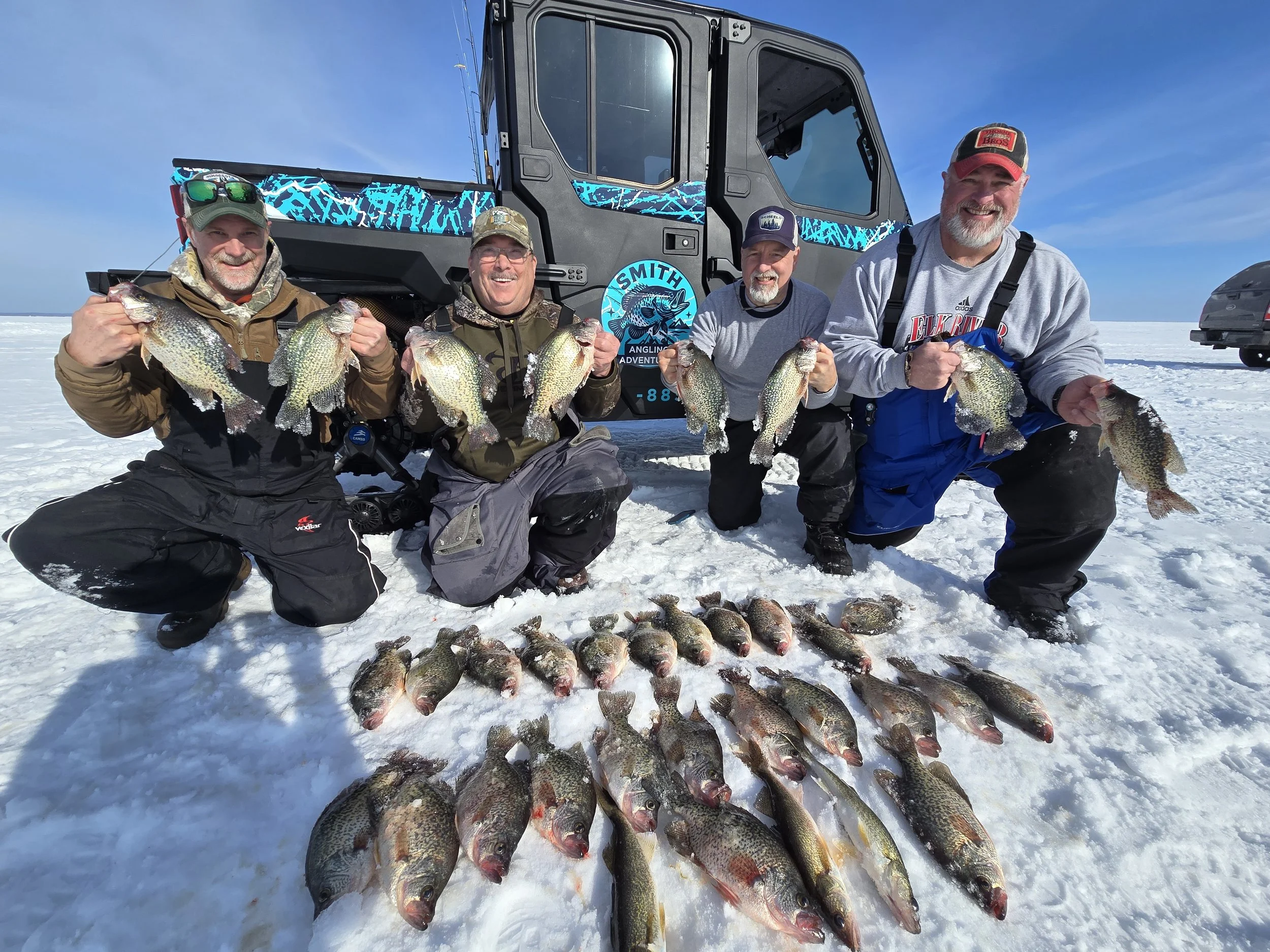 Four men on snow-covered ice holding fish and kneeling in front of a heavily loaded ice fishing vehicle, with fish laid out on the snow.
