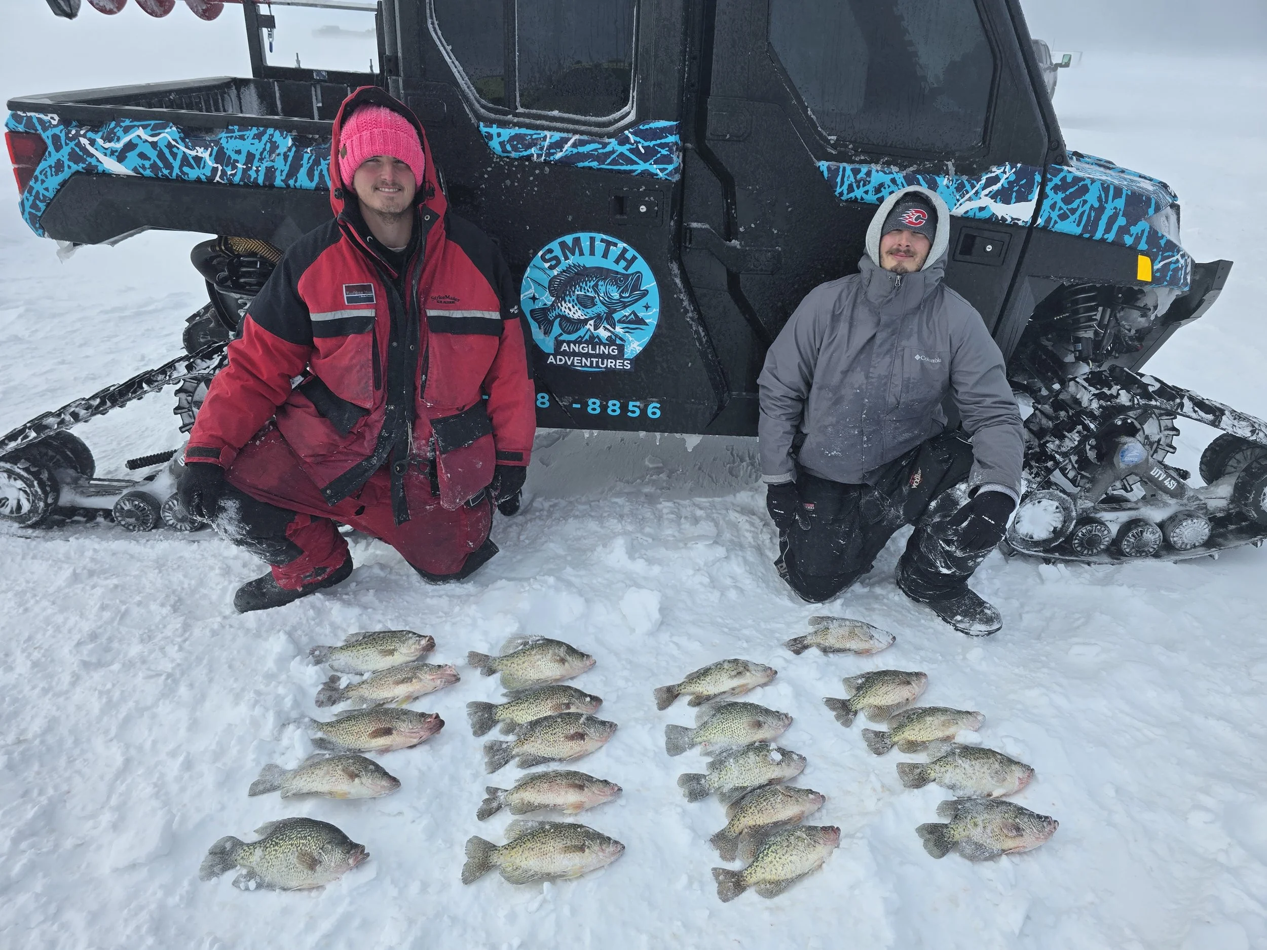 Two men in winter clothing kneel next to a row of caught fish on snow, with a snowmobile and a large off-road vehicle in the background.