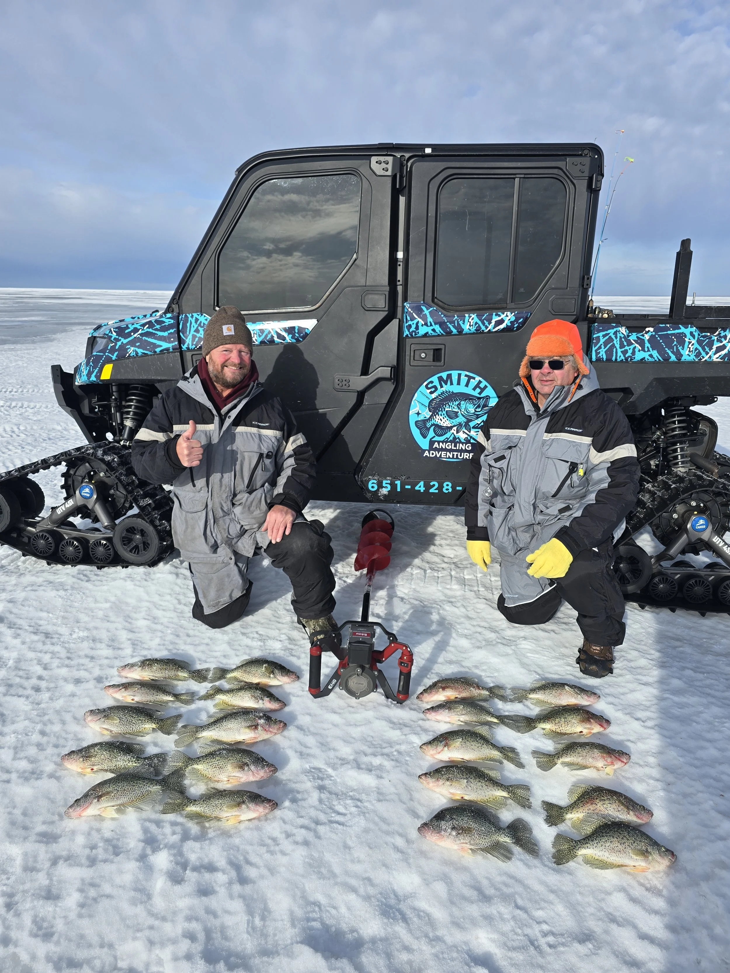 Two men in winter clothing kneeling on snow in front of a snowmobile with a team of fish caught from ice fishing, arranged on the snow.