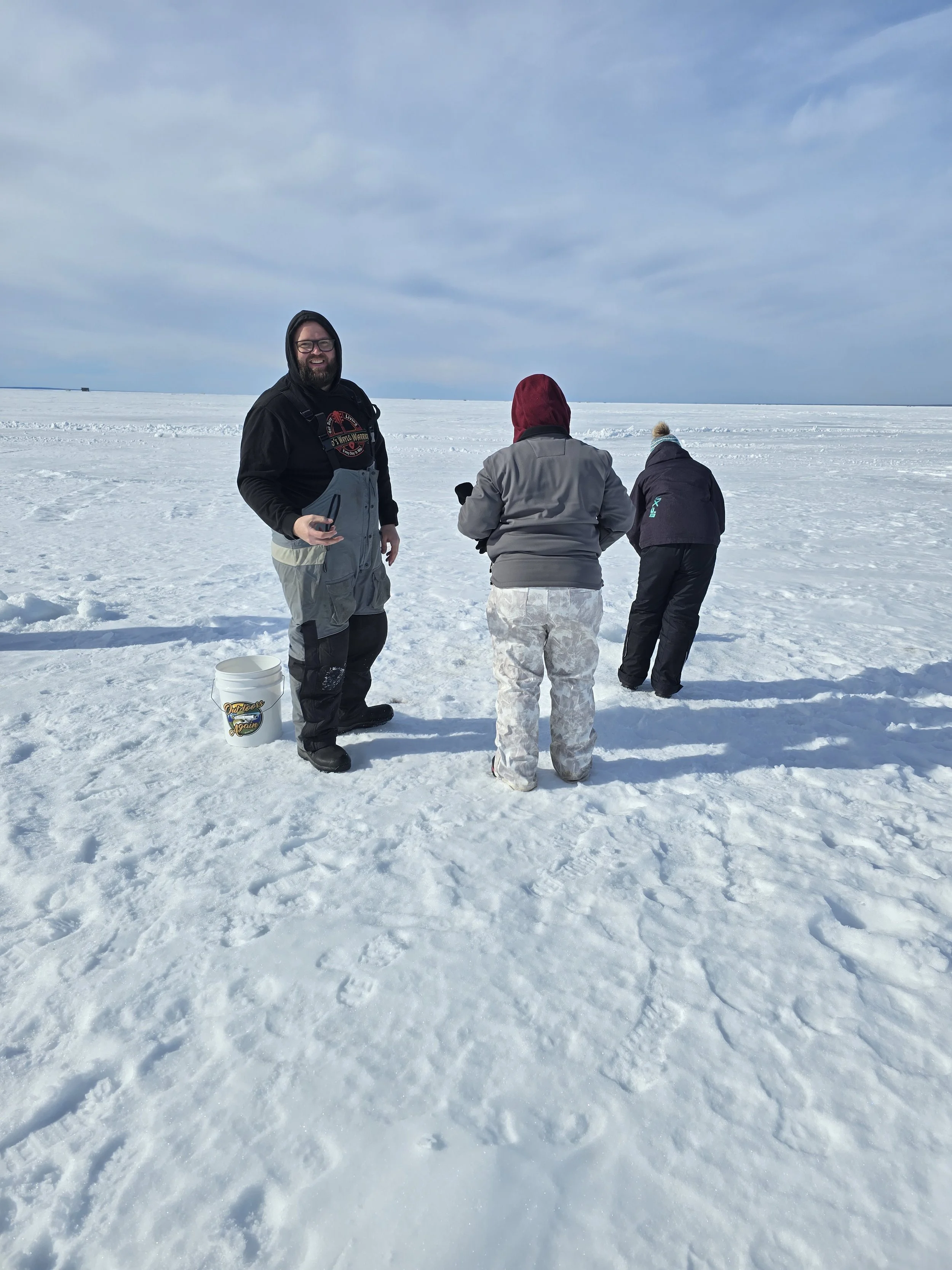 Three people stand on a snow-covered landscape under a partly cloudy sky, one smiling at the camera while the others face away.