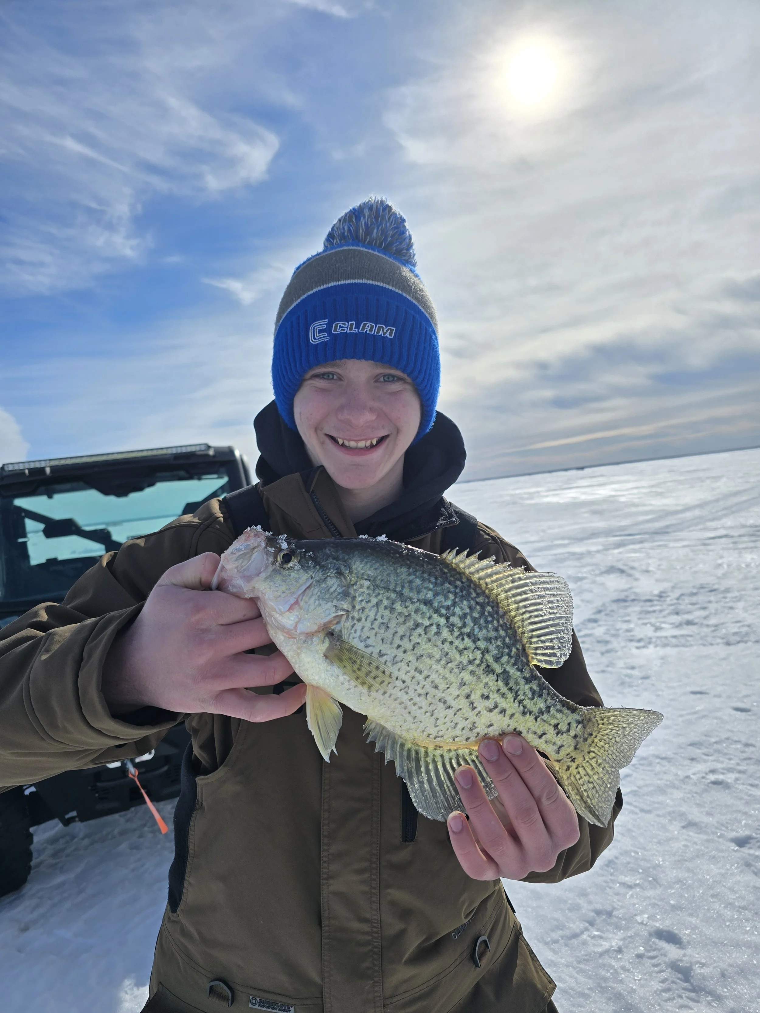 A person wearing a blue and gray beanie, smiling and holding a fish with yellow and green scales, on a snowy landscape with a cloudy sky and the sun in the background.