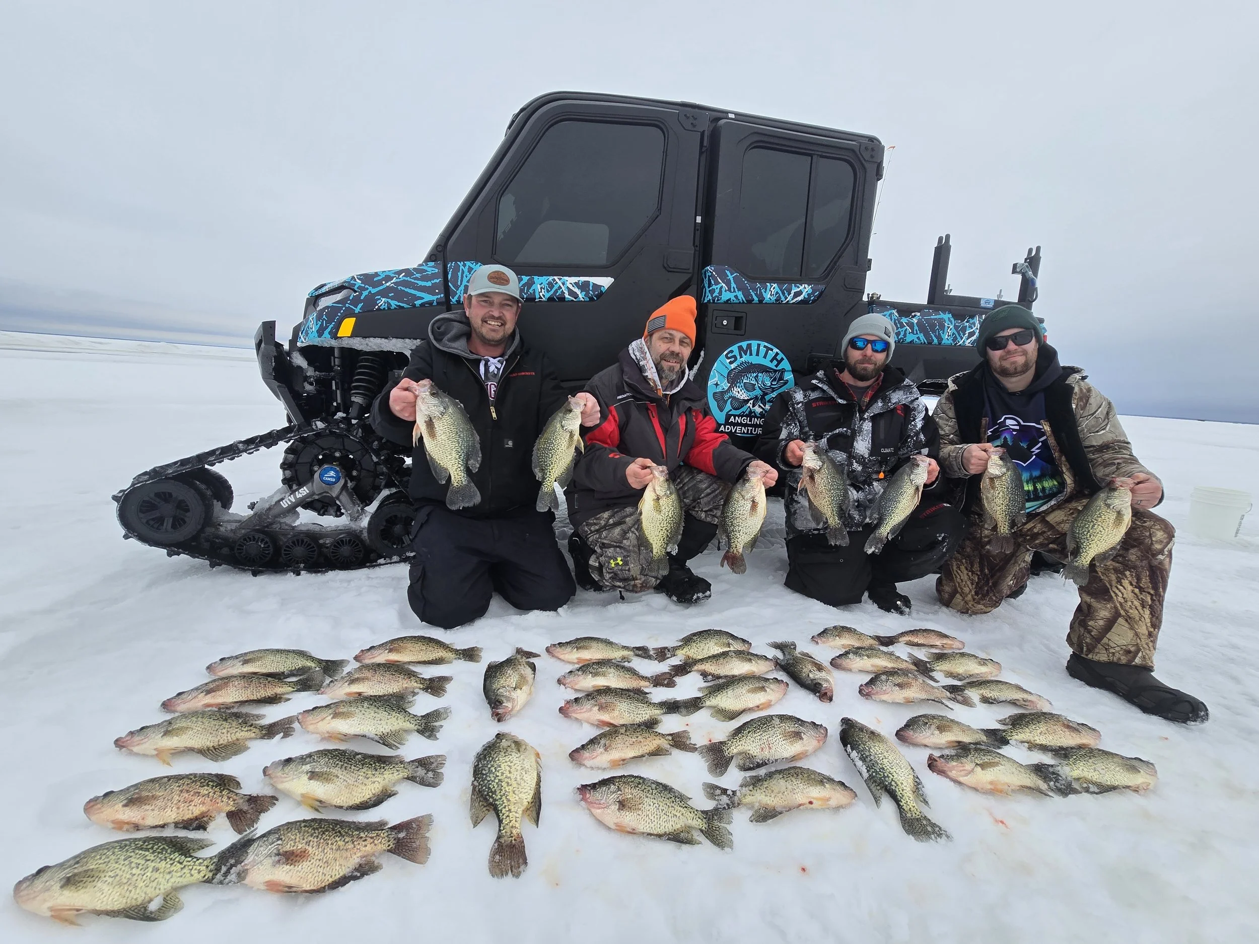 Four men kneeling in snow in front of a snowmobile, holding large fish they caught, with more fish arranged on the snow in front of them.