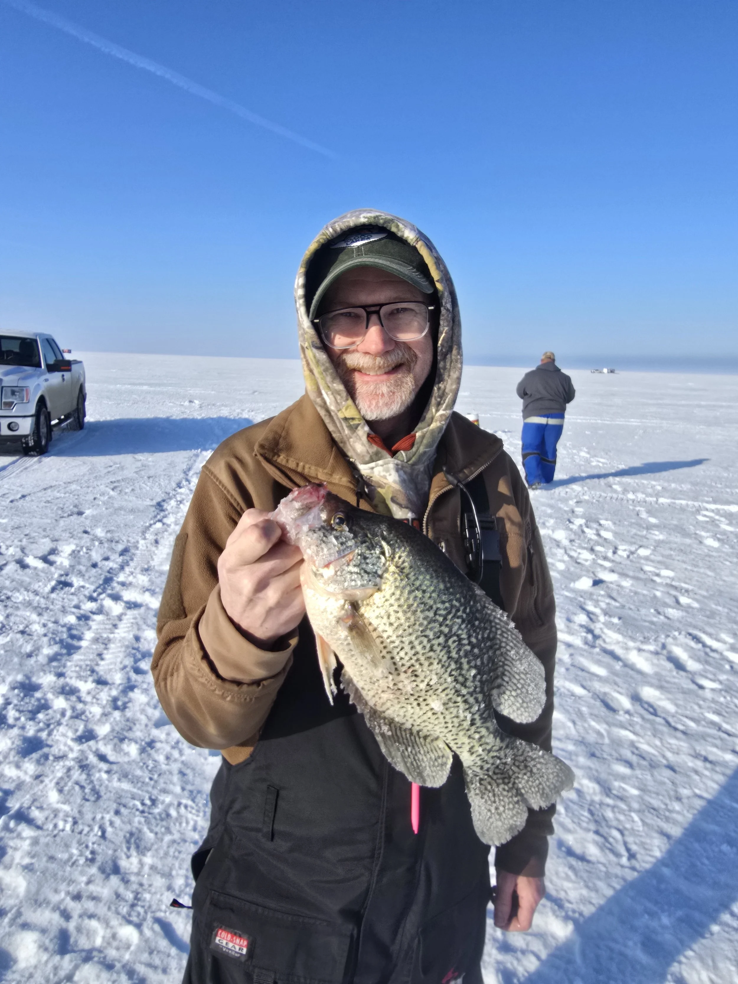 Man smiling while holding a fish on a snowy landscape, with a blue sky and a few people and vehicles in the background.