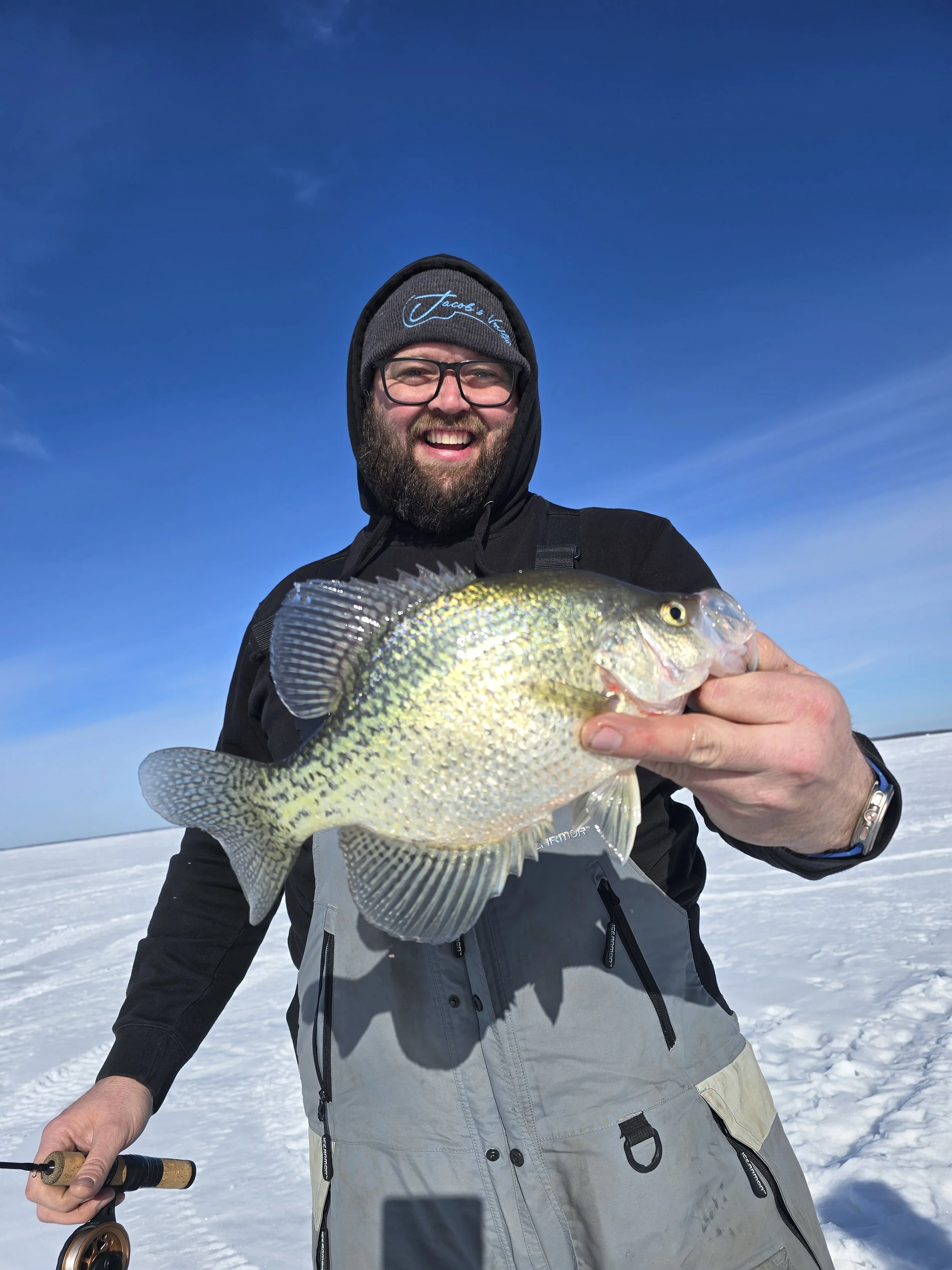A man with glasses, a beard, and a beanie hat smiling and holding a fish on a snowy landscape with a clear blue sky.