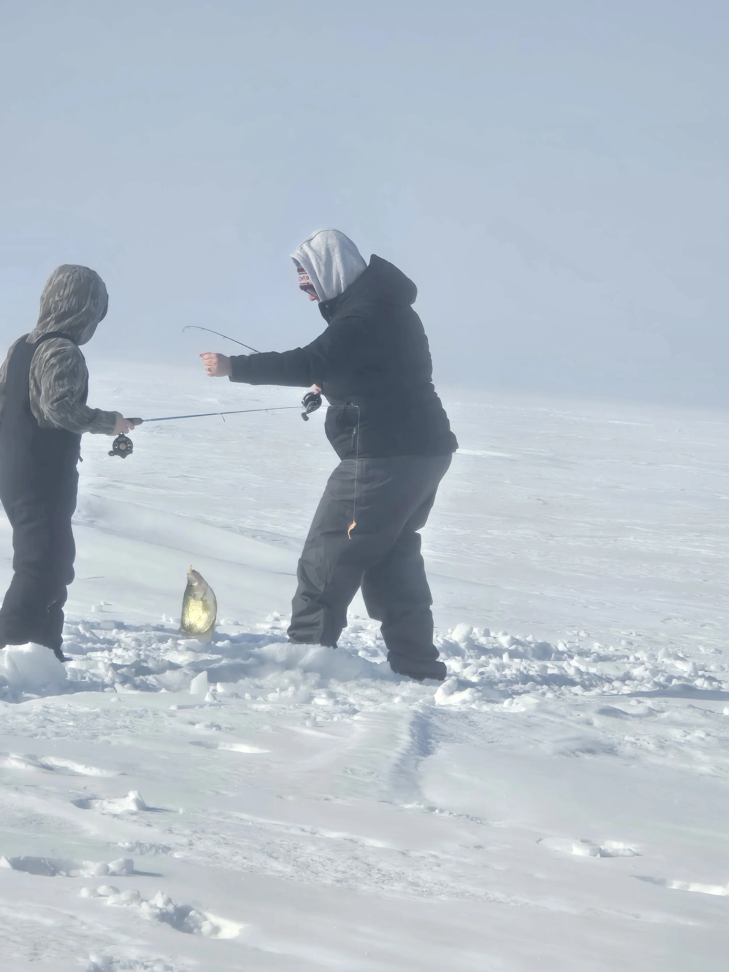 Two people ice fishing on a snowy, foggy landscape. One person is holding a fish, and the other is preparing or examining fishing equipment.