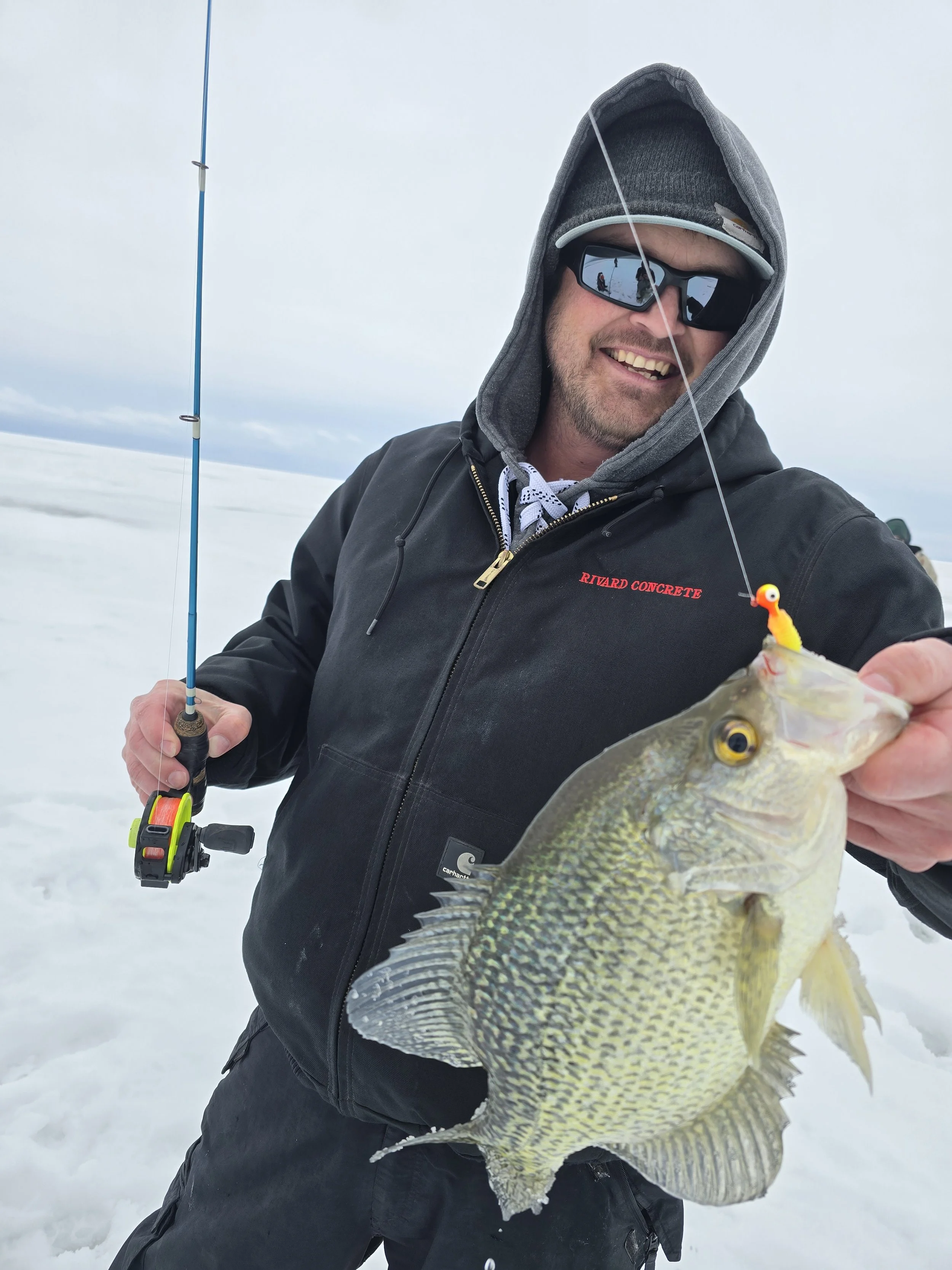 A man in winter clothing and sunglasses holding a fishing rod and displaying a large fish in a snowy landscape.