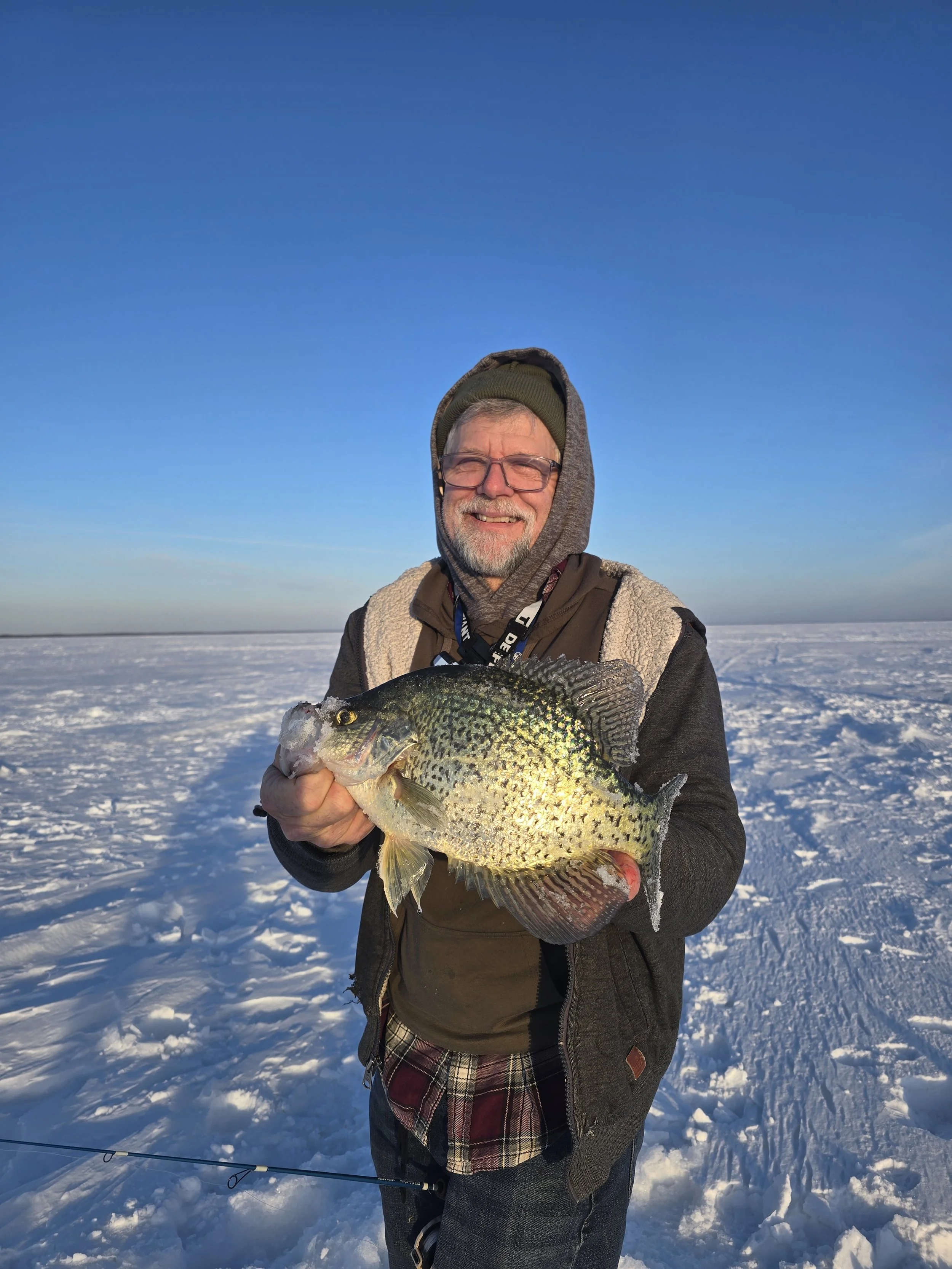 A man smiling and holding a large fish outdoors in a snowy landscape, dressed in winter clothing with sunglasses, under a clear blue sky.