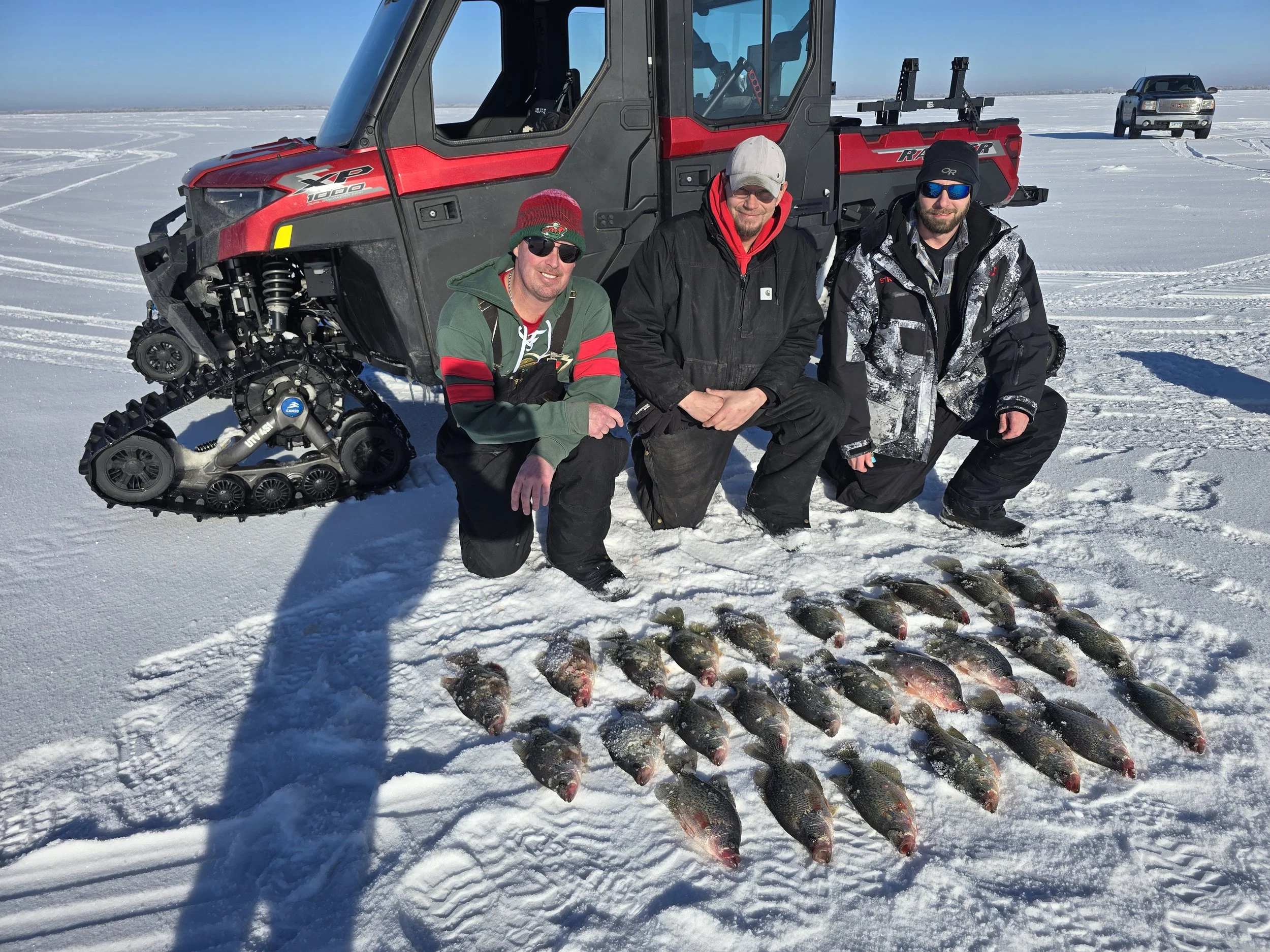 Three men in winter clothing kneeling on snow-covered ice in front of a line of fish caught from a frozen lake, with a red all-terrain vehicle and a pickup truck in the background.
