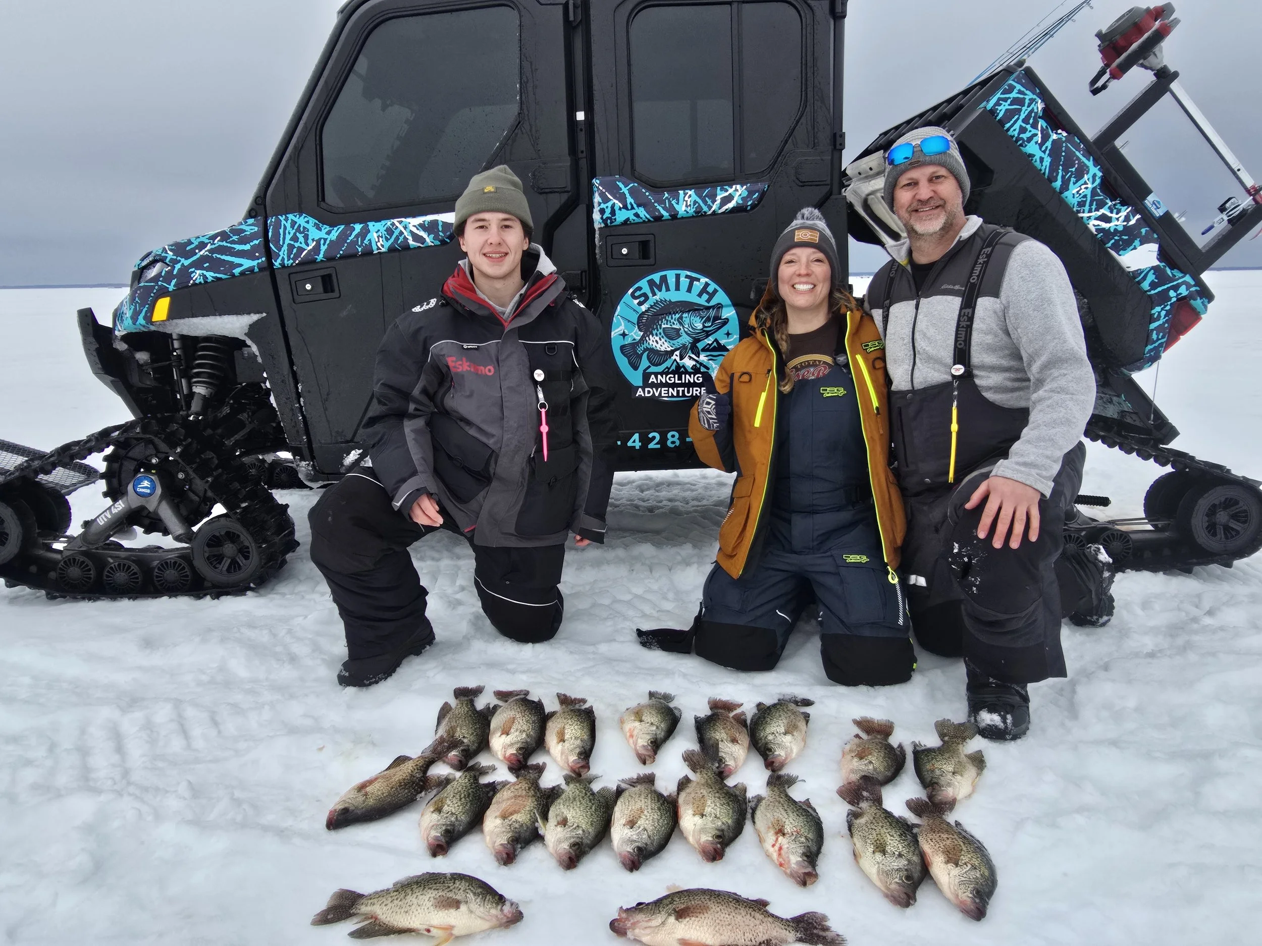 A group of three people dressed in winter outdoor clothing, smiling and posing behind a row of large freshly caught fish on snow, with a snow-covered landscape and overcast sky in the background, and a snowmobile with a marked vehicle nearby.