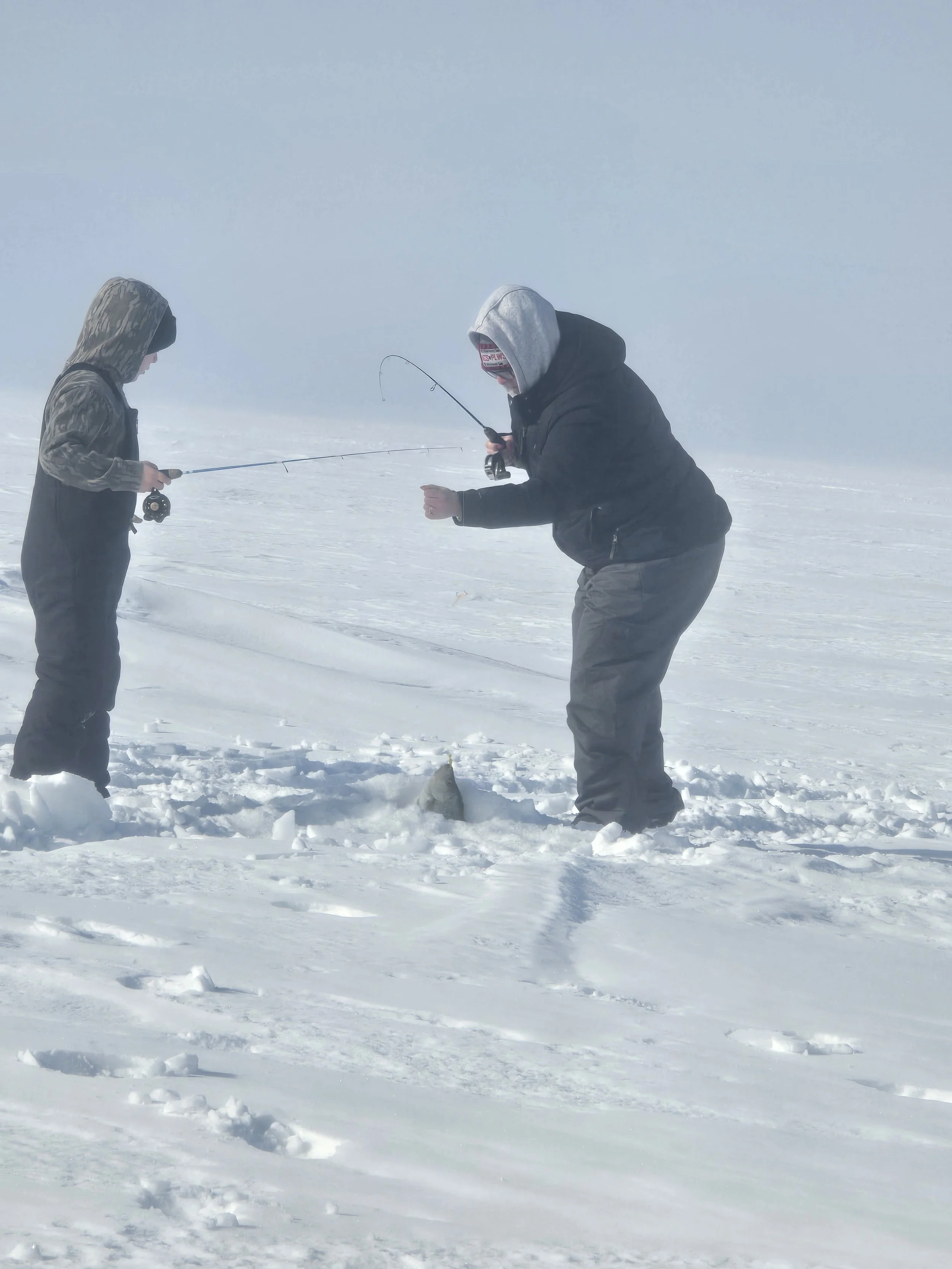 Two people in winter clothing are fishing on a snowy, foggy landscape.
