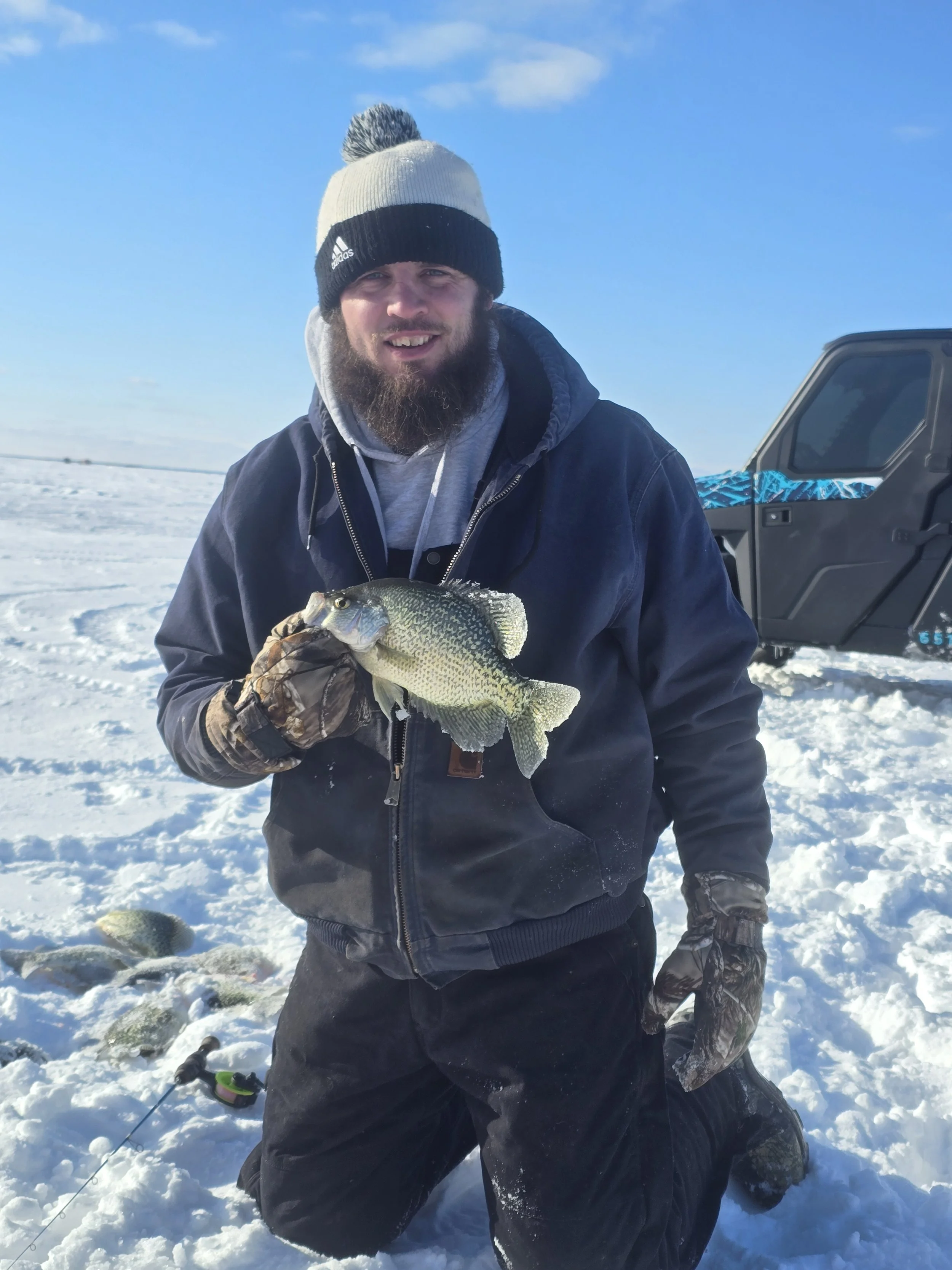 A man in winter clothing holding a fish on snowy ground with a snow-covered landscape and a blue sky in the background.