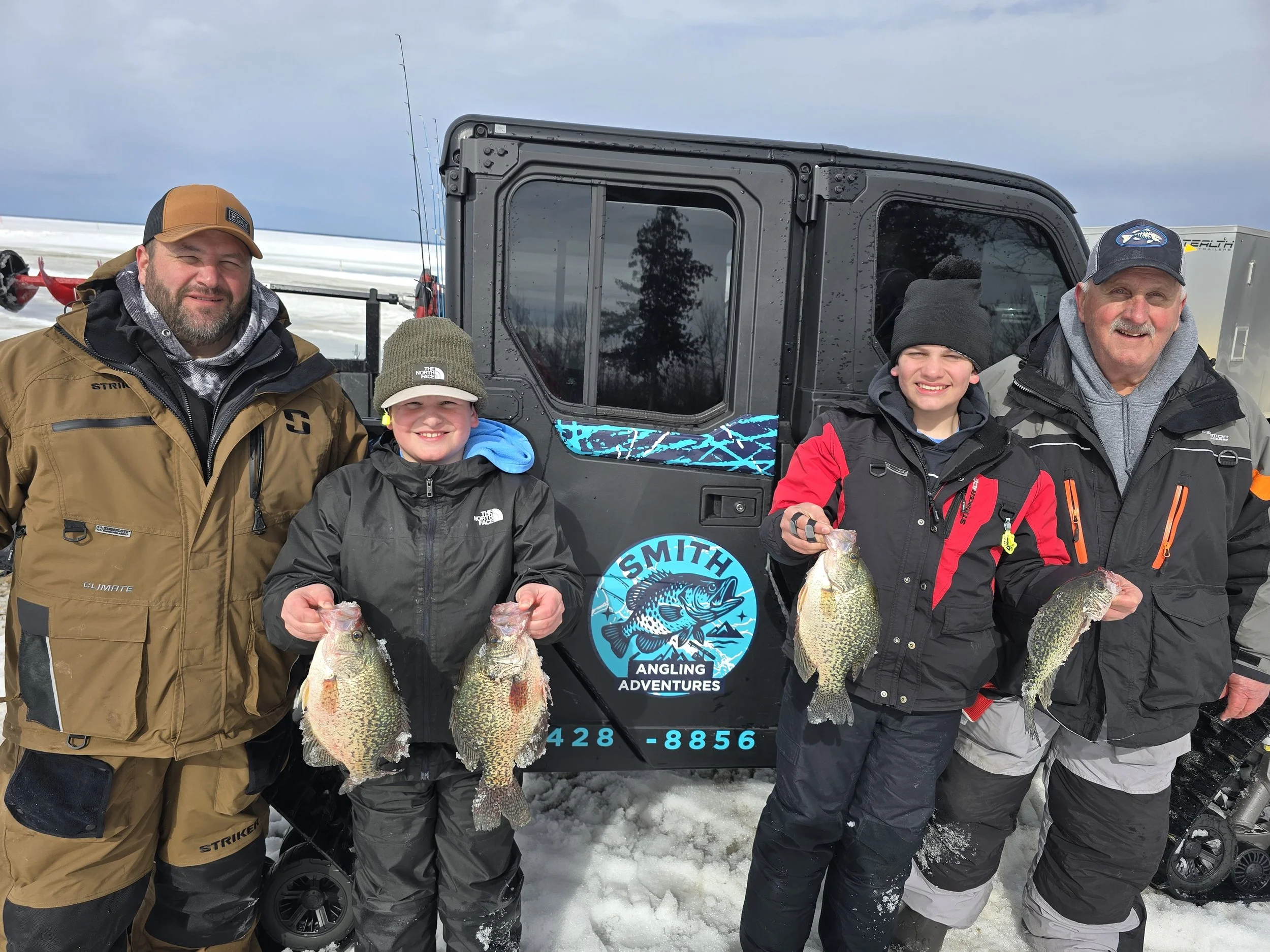 Four individuals, two adults and two children, posing outdoors in winter, each holding a large fish, standing in front of a black ATV with a sticker that reads "SMITH ANGLING ADVENTURES" and a phone number, on a snowy landscape with cloudy skies.