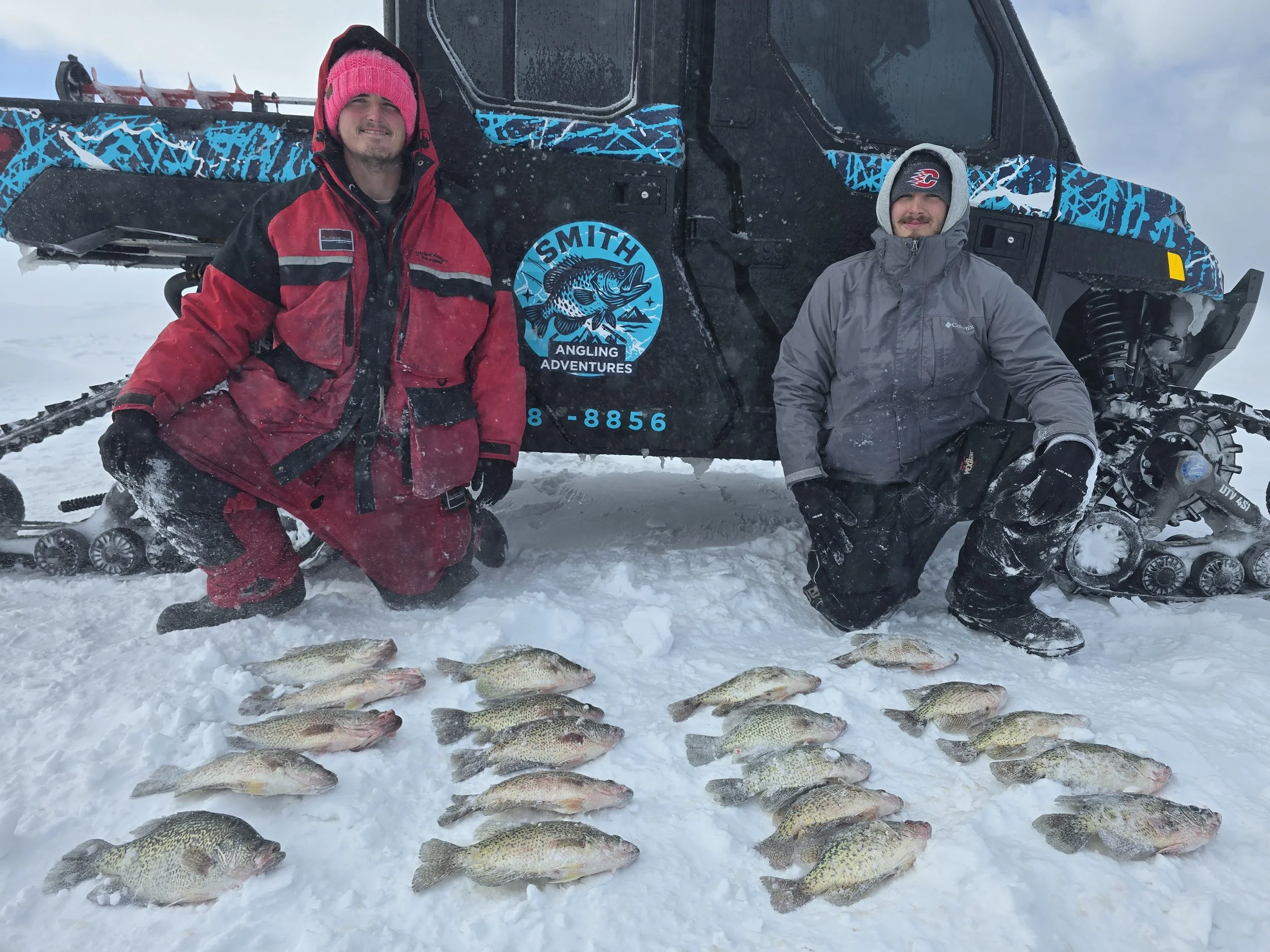 Two men in winter gear kneeling on snow next to a snowmobile with a group of fish laid out on the snow in front of them.