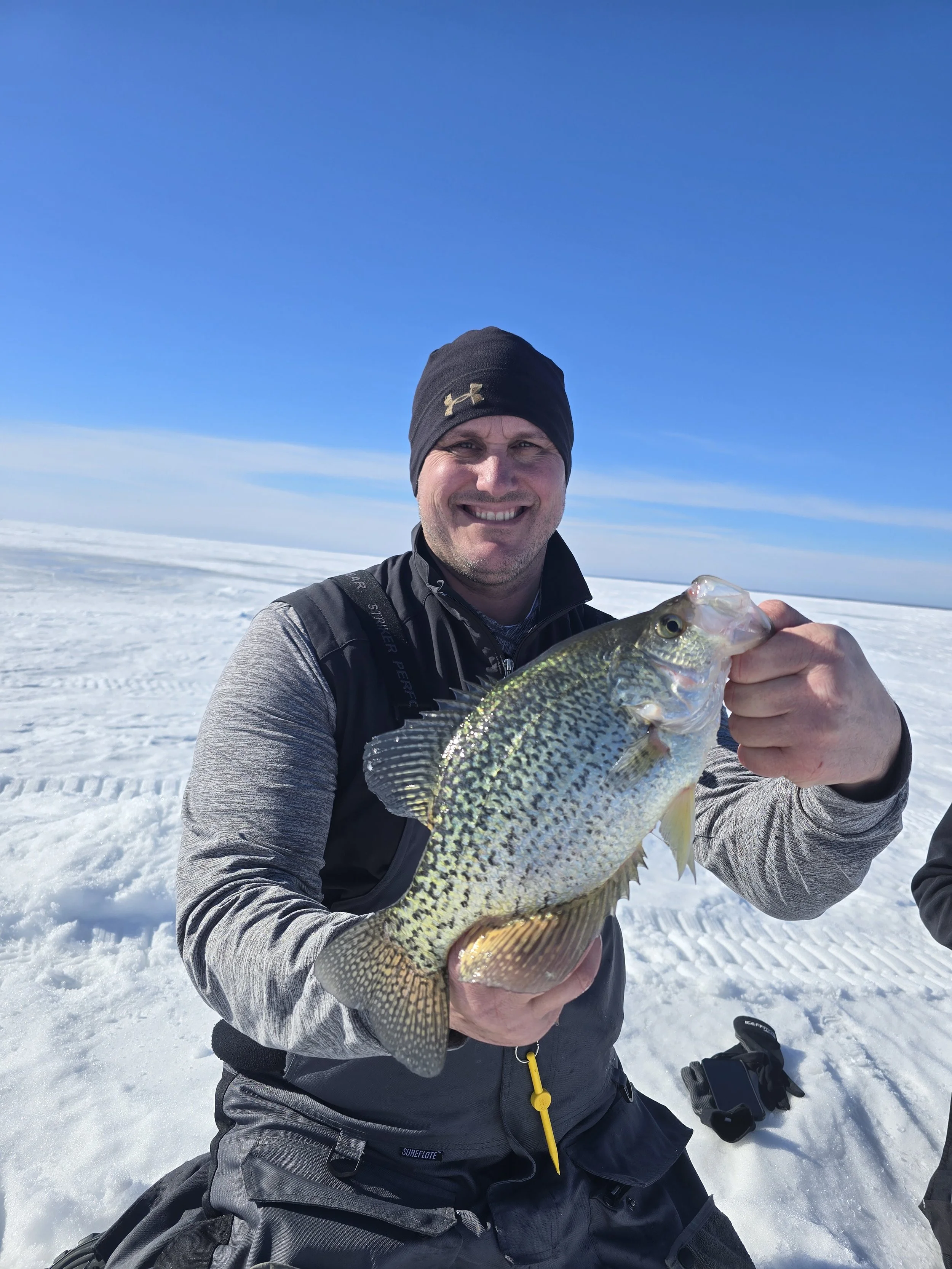 Man dressed in winter clothing holding a fish on snow-covered ground in a snowy landscape under blue sky.