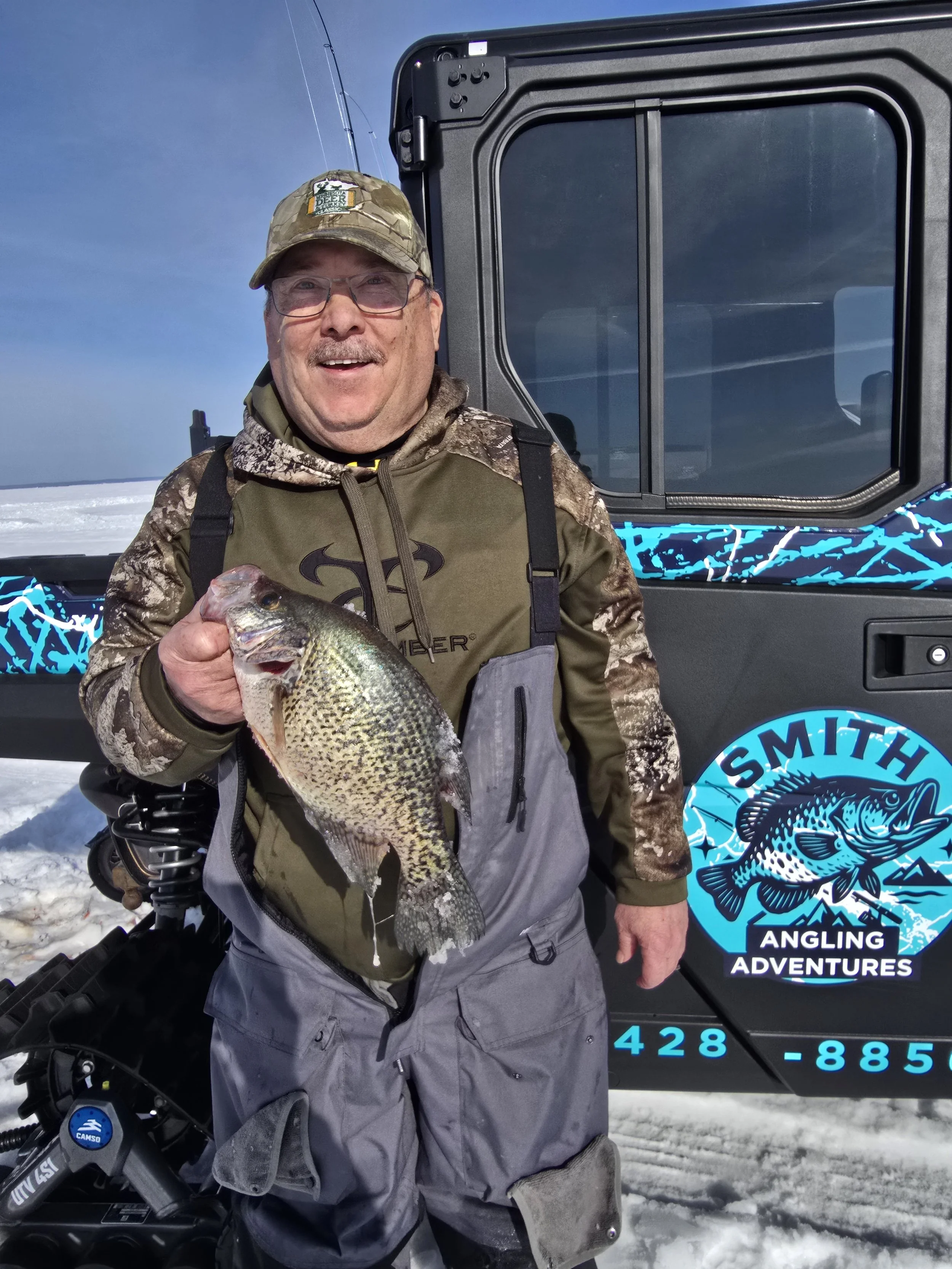 A man in hunting gear holding a large fish, standing in snowy terrain with a vehicle in the background that displays a logo for 'Smith Angling Adventures'.