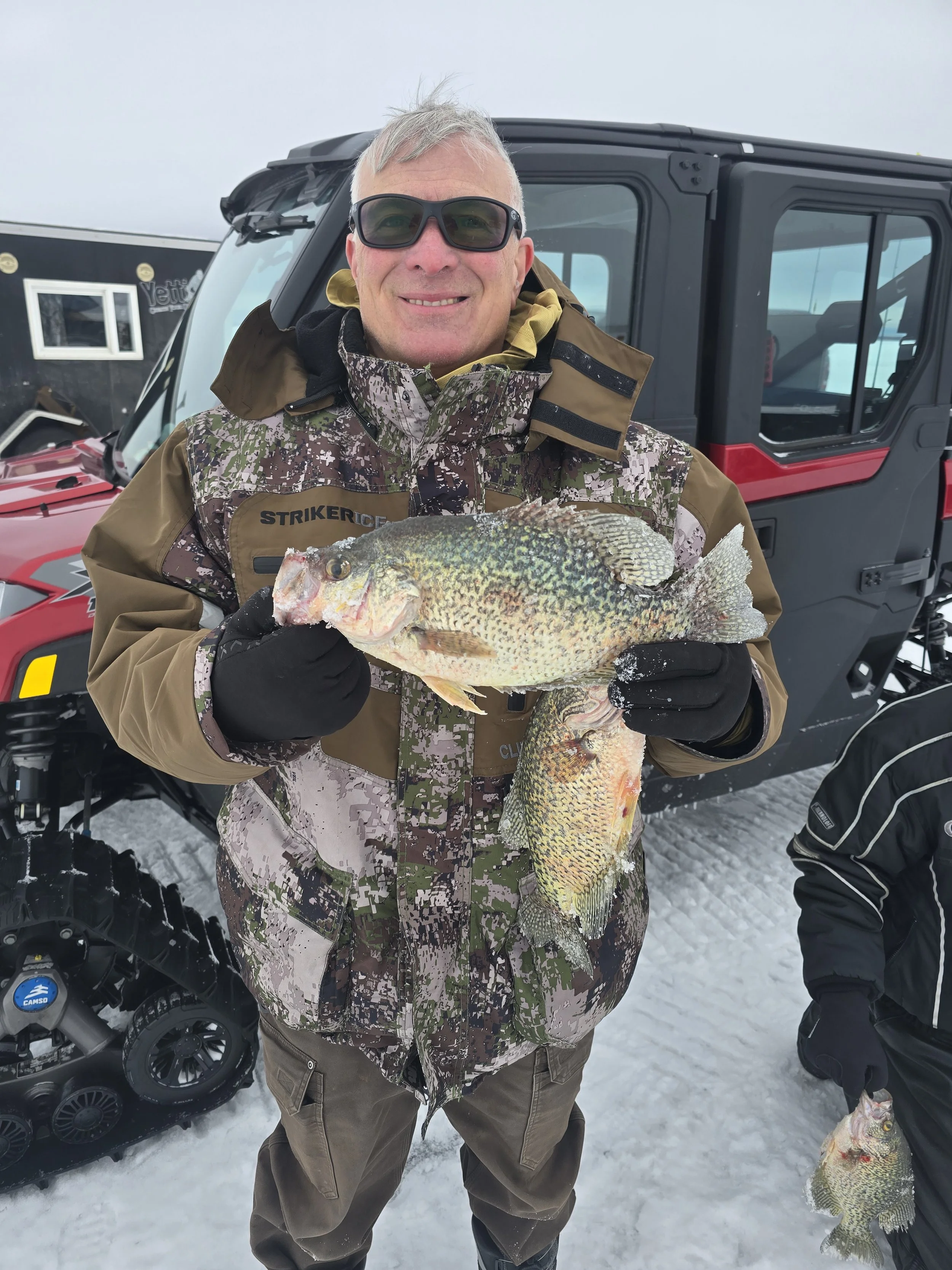 A man with gray hair wearing sunglasses, a camouflage and brown winter jacket, and brown pants, is smiling and holding a large fish with yellow and green scales, in a snowy outdoor setting with a red and black ATV and a black vehicle in the backgroun