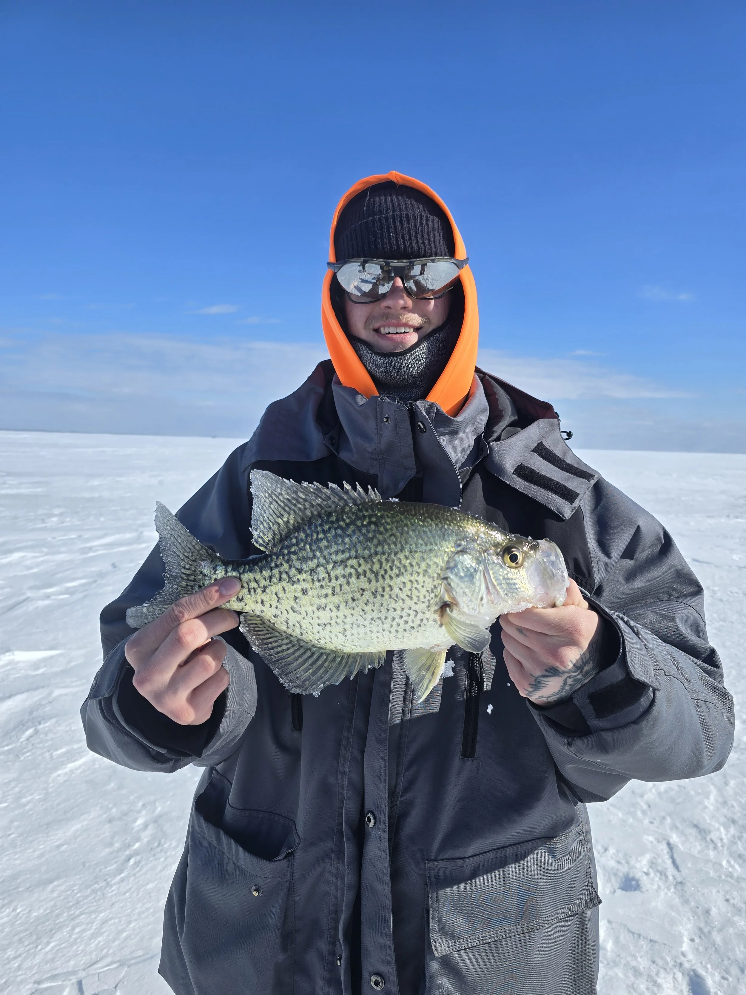 A person dressed in winter gear holding a fish on a snowy, flat landscape with a clear blue sky.