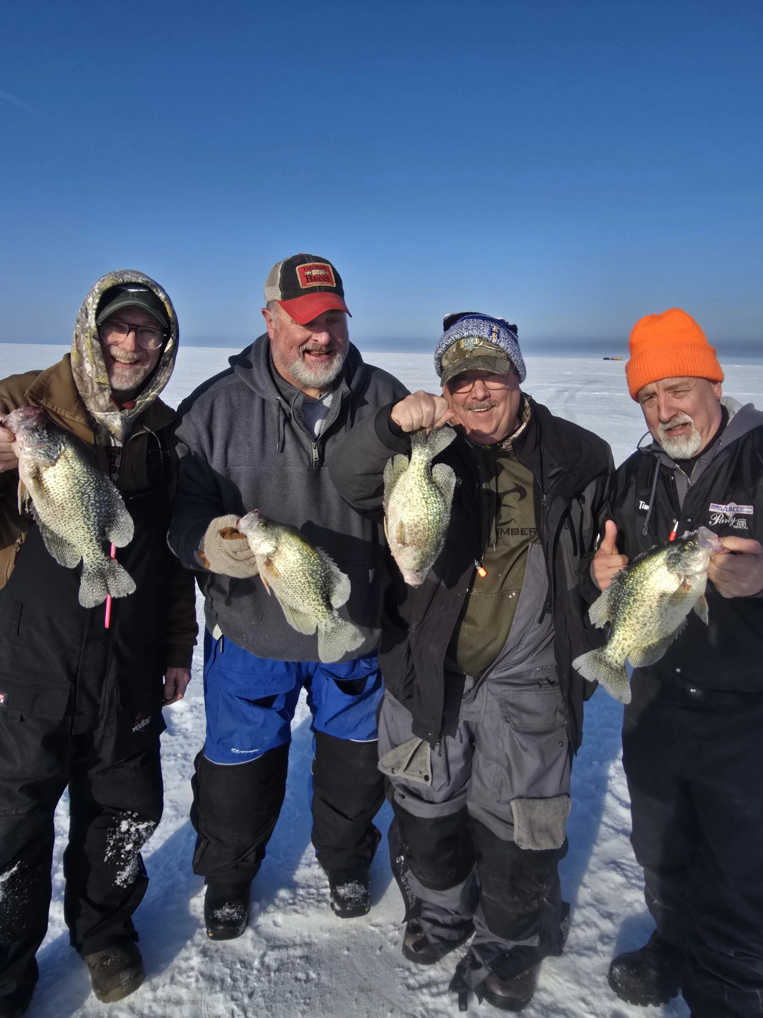Four men in winter clothing standing on snow and ice, holding fish, smiling, and posing for the photo, with a clear blue sky in the background.