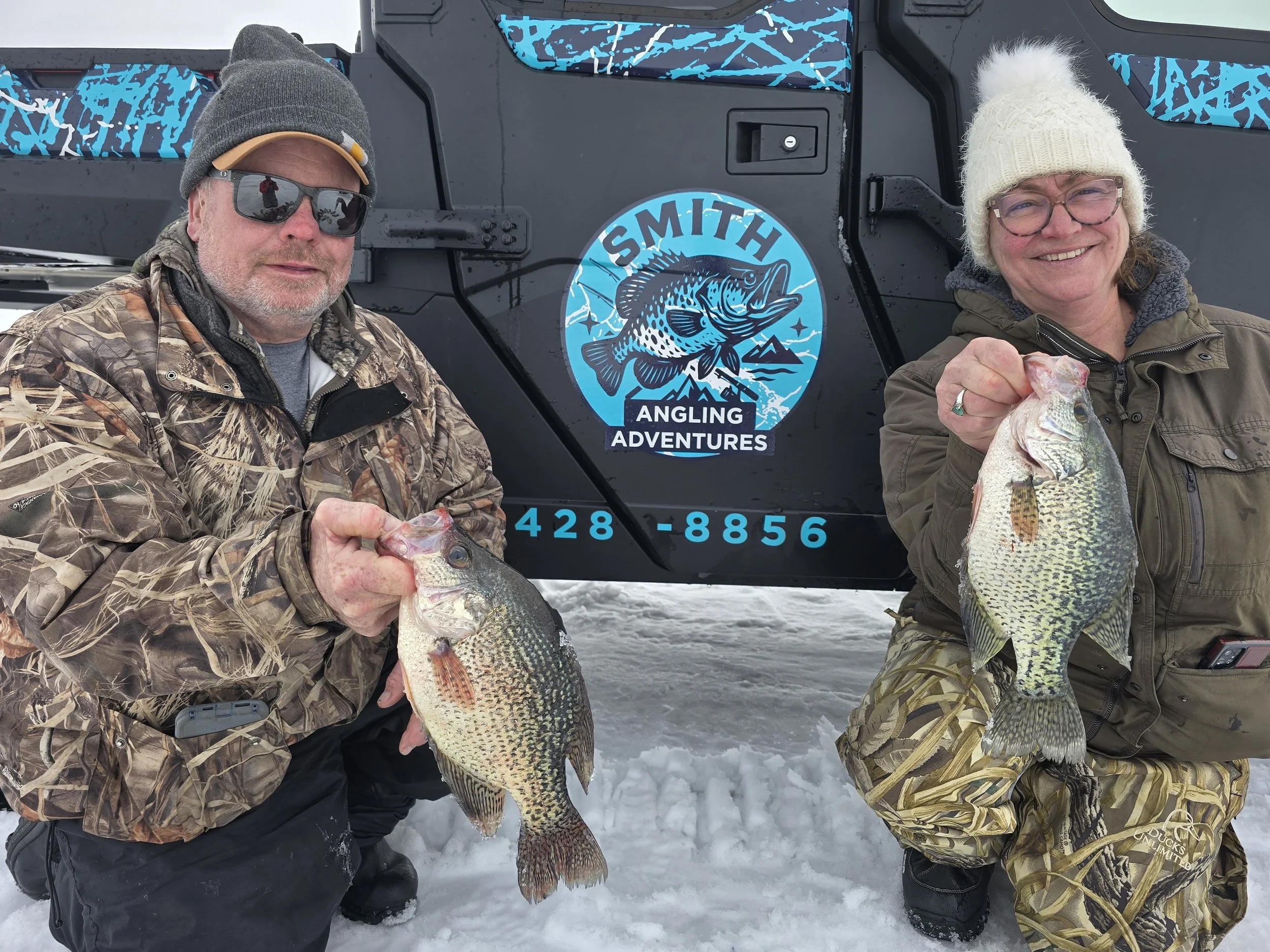 Two people dressed in winter clothing holding large fish they caught in the snow, standing in front of a vehicle with a blue logo that reads 'Smith Angling Adventures'.