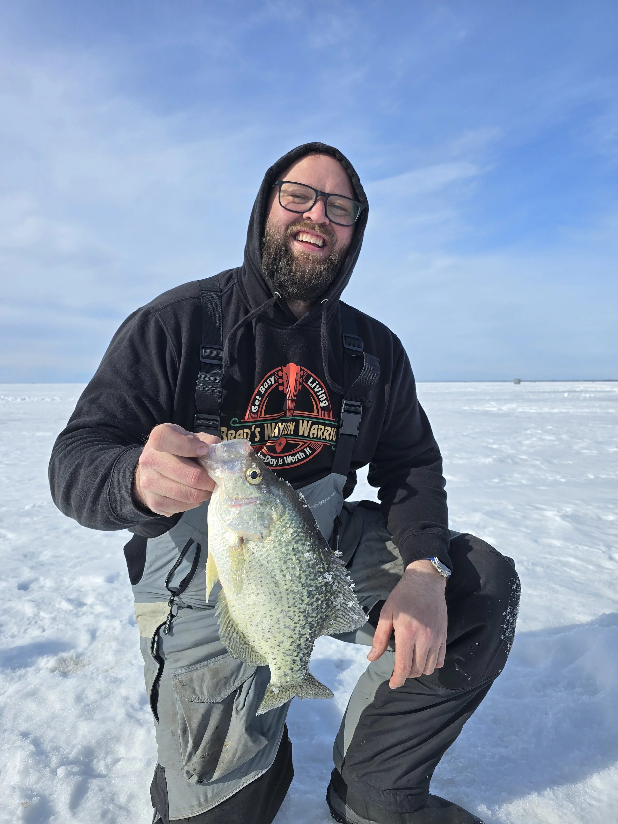 A man with glasses and a beard dressed in winter clothing is kneeling on snow, holding a large fish he caught, with a blue sky in the background.