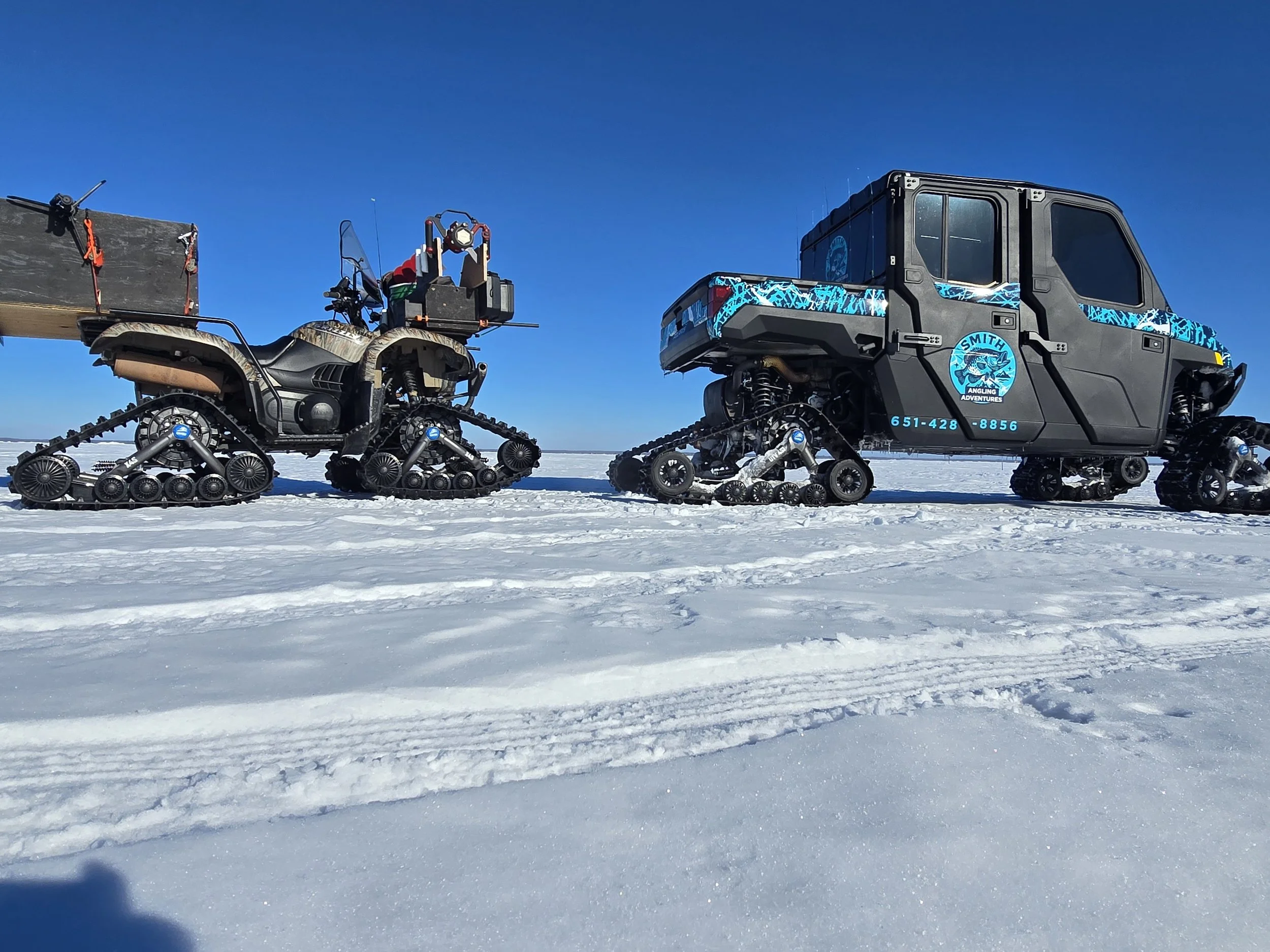Snow-covered landscape with two tracked snow vehicles, one with a box and one with a sign that says 'Smith Angling Adventures', under a clear blue sky.